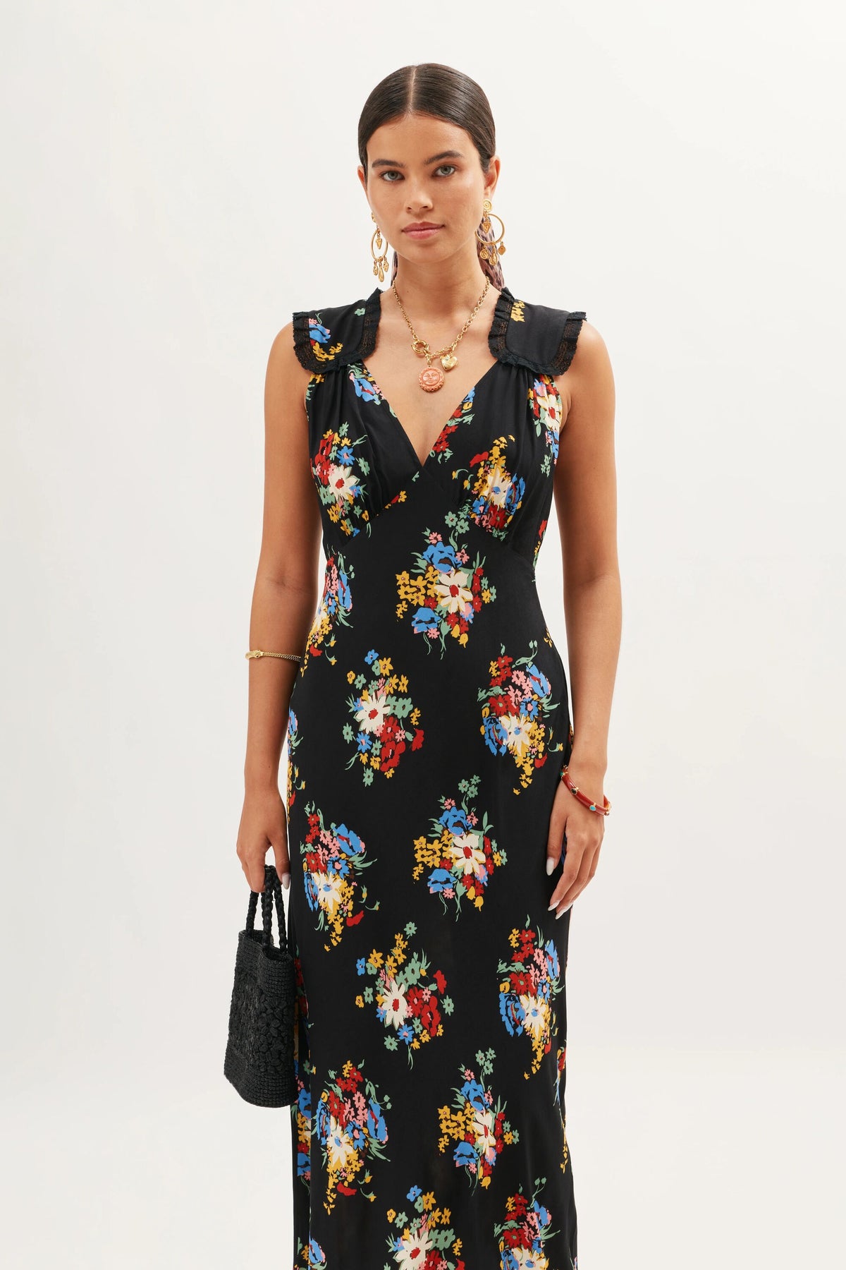 A woman wears the Chessie Darcie Floral Black dress, a sleeveless black piece featuring a vibrant floral print, ruffled shoulders, and a deep V-neckline. She styles it with gold jewelry and carries a black textured handbag against a simple background.