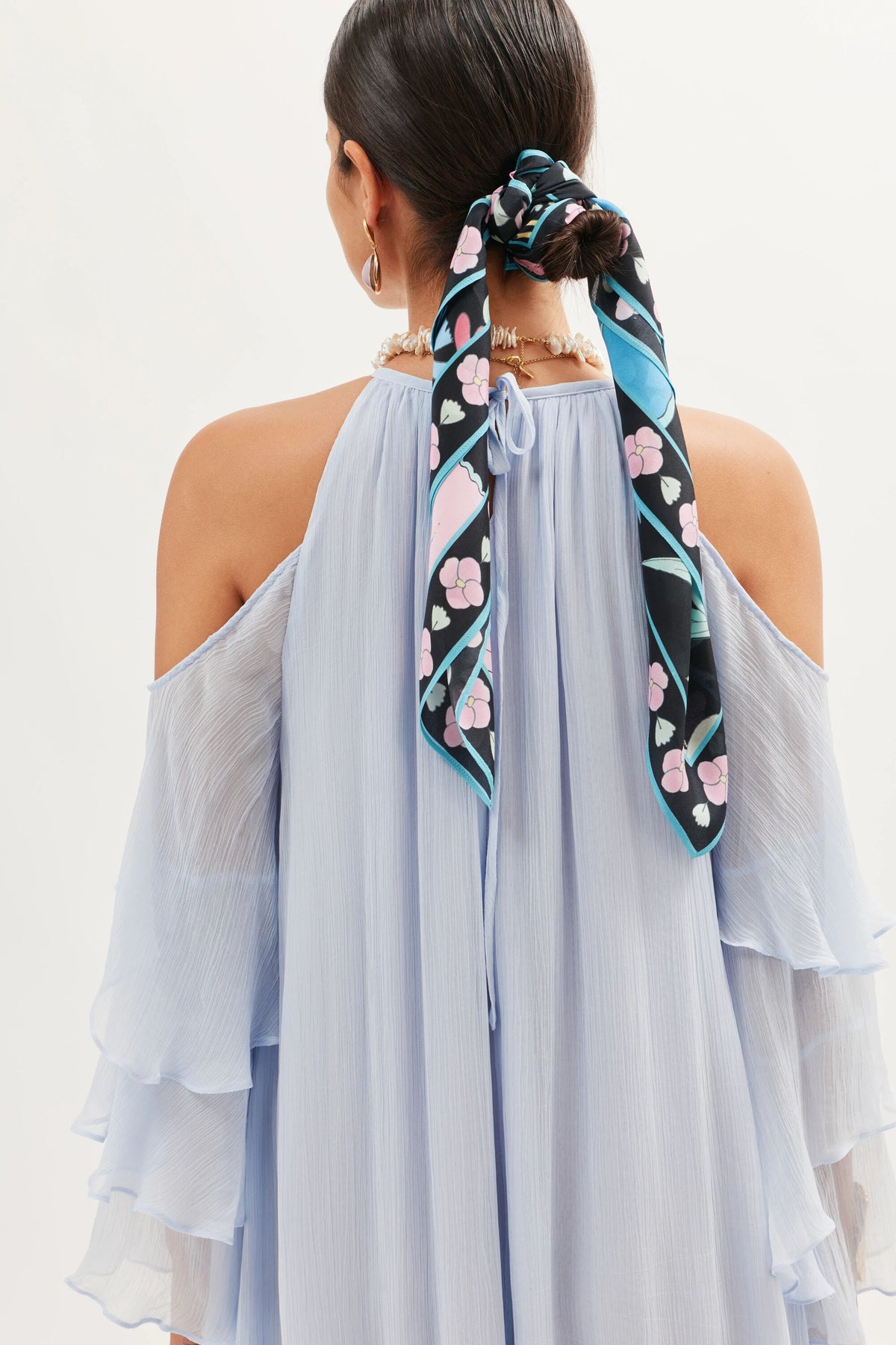 A woman with dark hair in a low ponytail tied with a black floral scarf models the Niara – Periwinkle gown, featuring off-the-shoulder ruffles, as seen from the back against a plain background.
