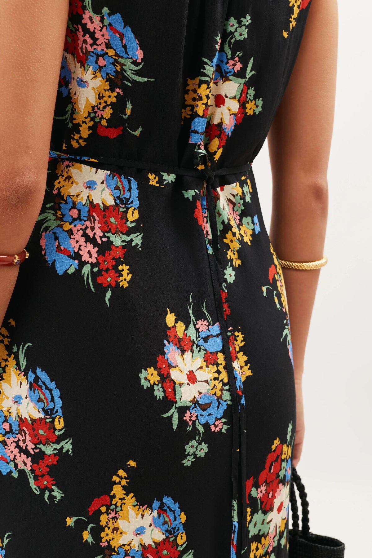 A close-up of a person wearing the Chessie - Darcie Floral Black dress, featuring colorful floral prints. The person accessorizes with a gold bracelet and holds a black woven bag, with the image highlighting the back and waist area of the dress.