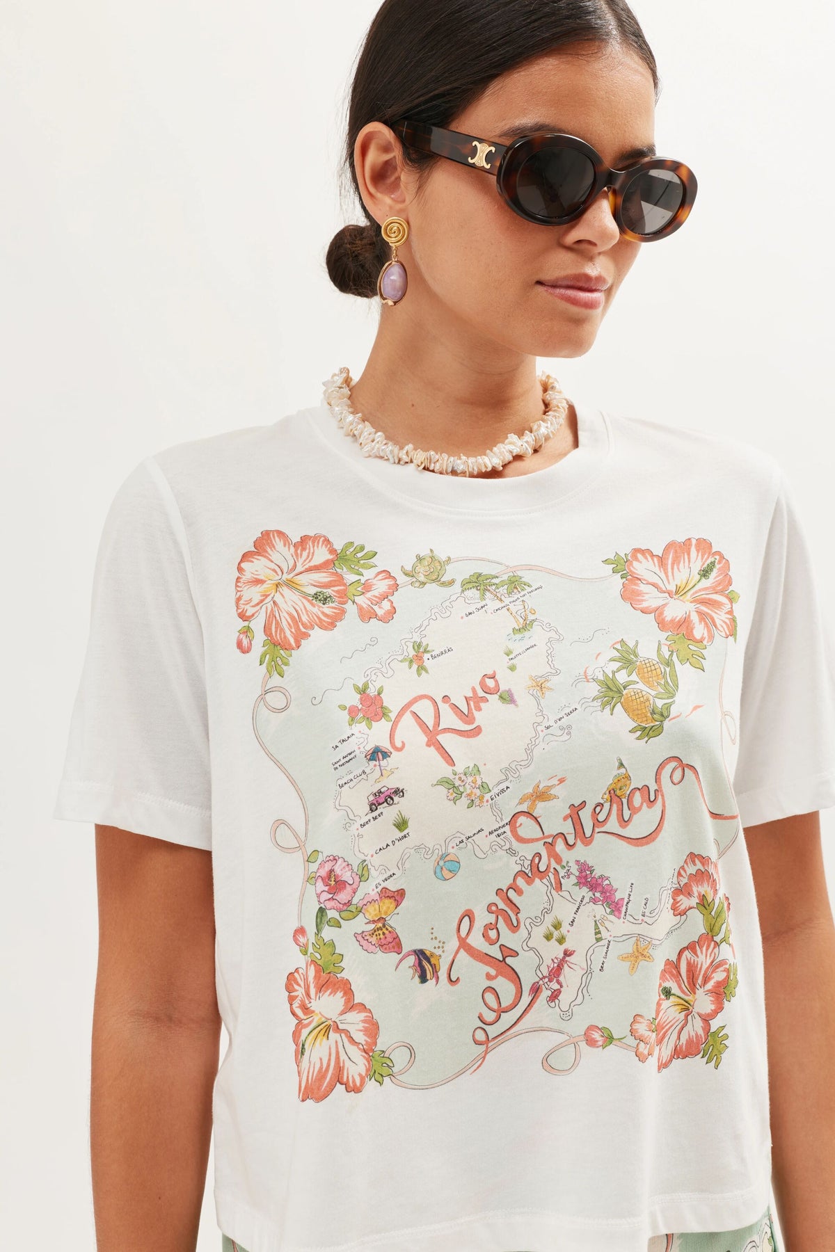 A woman wears oversized sunglasses, drop earrings, and a Sabria - Ibiza Souvenir White T-shirt with Riva Ventura in colorful script and tropical flowers. Her hair is tied back with a seashell necklace for a vintage poster vibe.