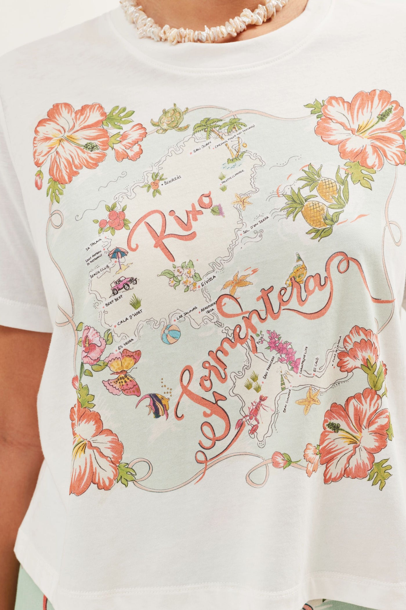 The Sabria - Ibiza Souvenir White T-shirt features a vintage-style Ibiza map with Río and Formentera, adorned with flowers, fruit, landmarks, and large orange hibiscus blooms.