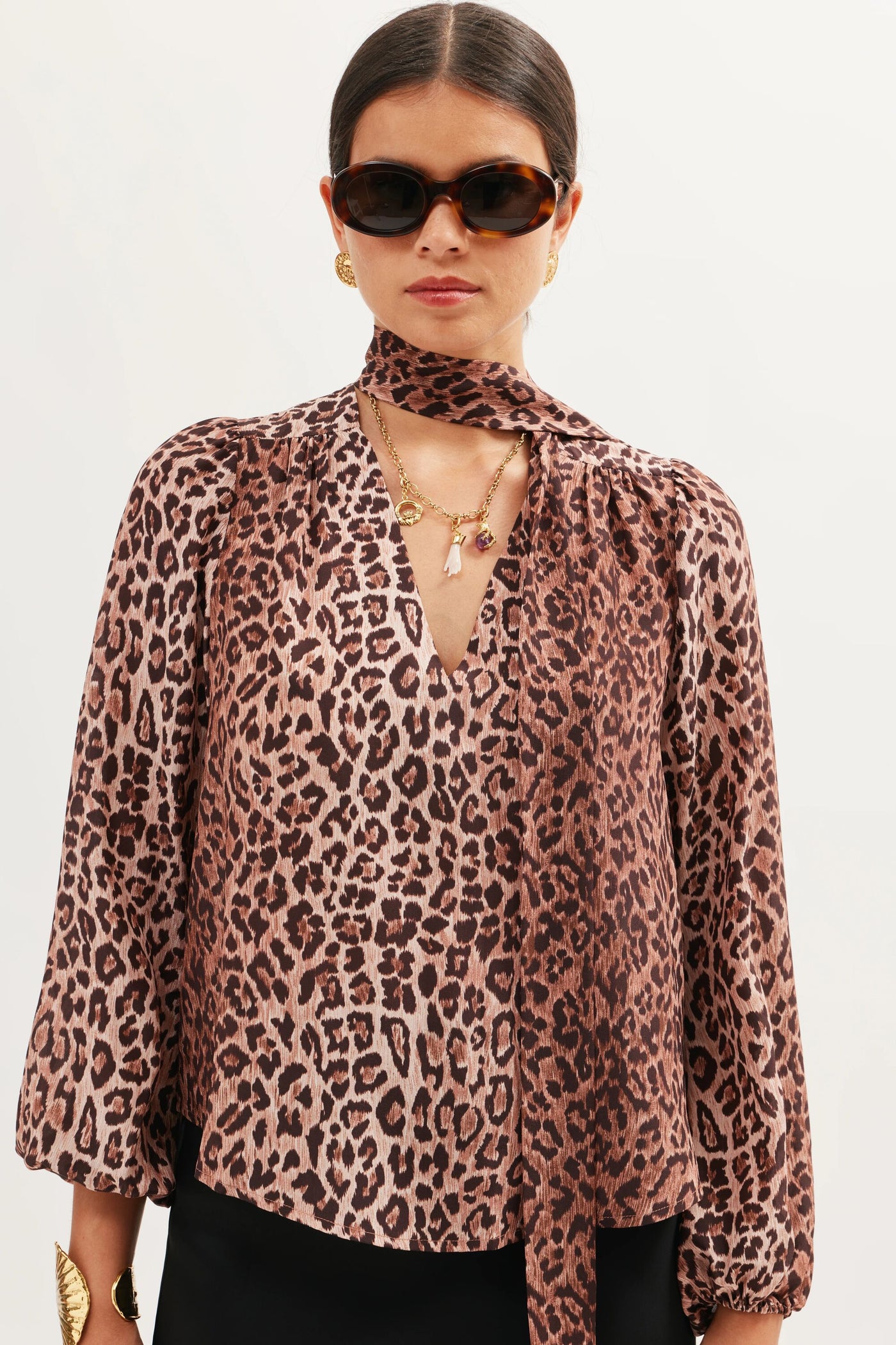 A woman with dark hair tied back wears sunglasses and the Lorene - Gold necklace, paired with a brown leopard-print blouse, matching scarf tie, and black pants, standing against a plain white background.