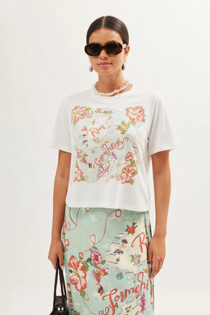 A woman in dark sunglasses, a beaded necklace, and the Sabria - Ibiza Souvenir White T-shirt with colorful floral script designs, paired with a matching floral skirt, stands against a light background evoking vintage poster vibes.