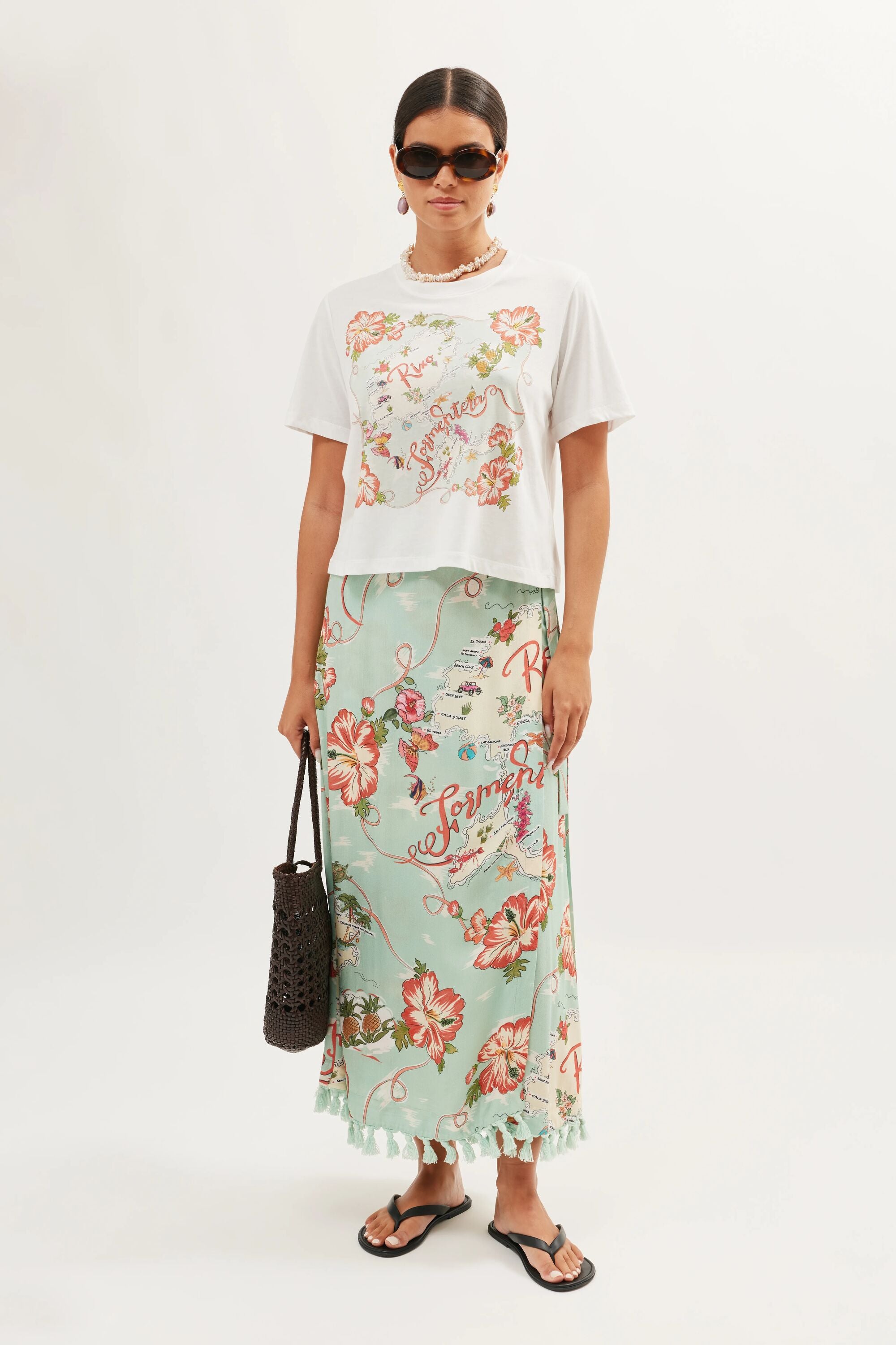 A woman in sunglasses, a white Sabria - Ibiza Souvenir White T-shirt with floral print, a long light blue floral skirt, black flip-flops, and holding a dark woven bag stands against a plain light background.