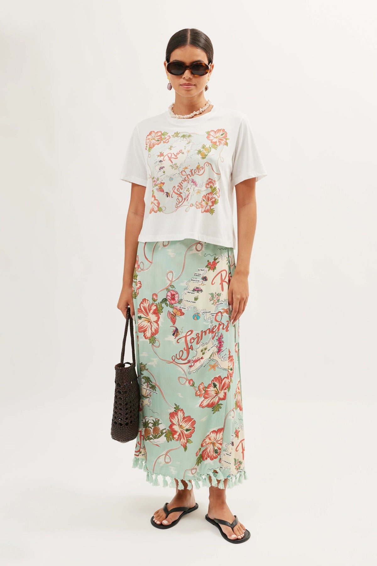A woman in sunglasses, a white Sabria - Ibiza Souvenir White T-shirt with floral print, a long light blue floral skirt, black flip-flops, and holding a dark woven bag stands against a plain light background.