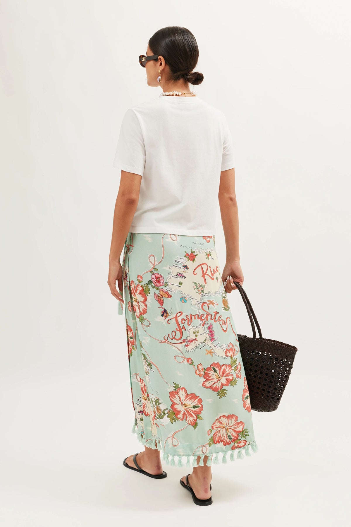 A woman with dark hair in a low bun wears sunglasses, the Sabria Ibiza Souvenir White T-shirt, a light green floral wrap skirt with red and pink flowers, black sandals, and carries a black woven tote against a plain background.