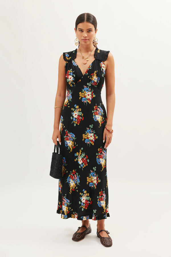A woman wearing the Chessie - Darcie Floral Black sleeveless dress with ruffled shoulders, paired with brown woven shoes, gold jewelry, and holding a black handbag stands against a plain white background.
