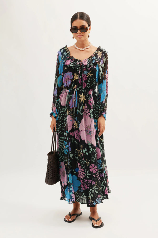 A woman in the Alina - Bhatia Floral Black long sleeve dress with boho vibes, accessorized with a beaded necklace, black woven bag, flip-flops, sunglasses, and her hair pulled back for an easygoing look.