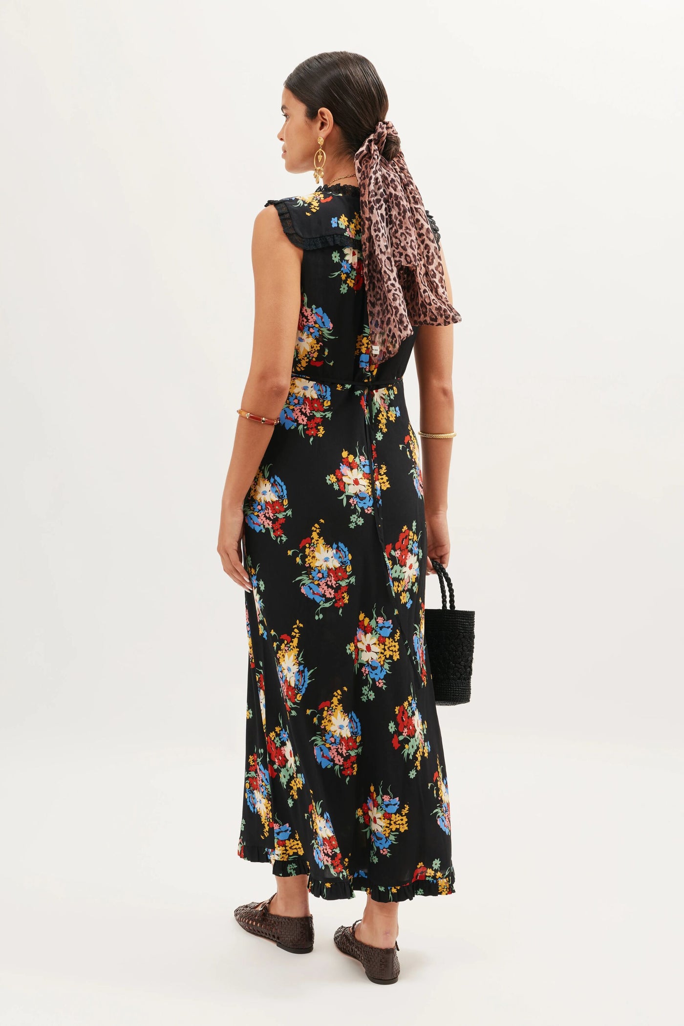 A woman stands facing away, wearing the Chessie - Darcie Floral Black sleeveless dress, paired with brown woven shoes, gold jewelry, and a leopard print scarf in her hair. She holds a black textured handbag against a plain white background.
