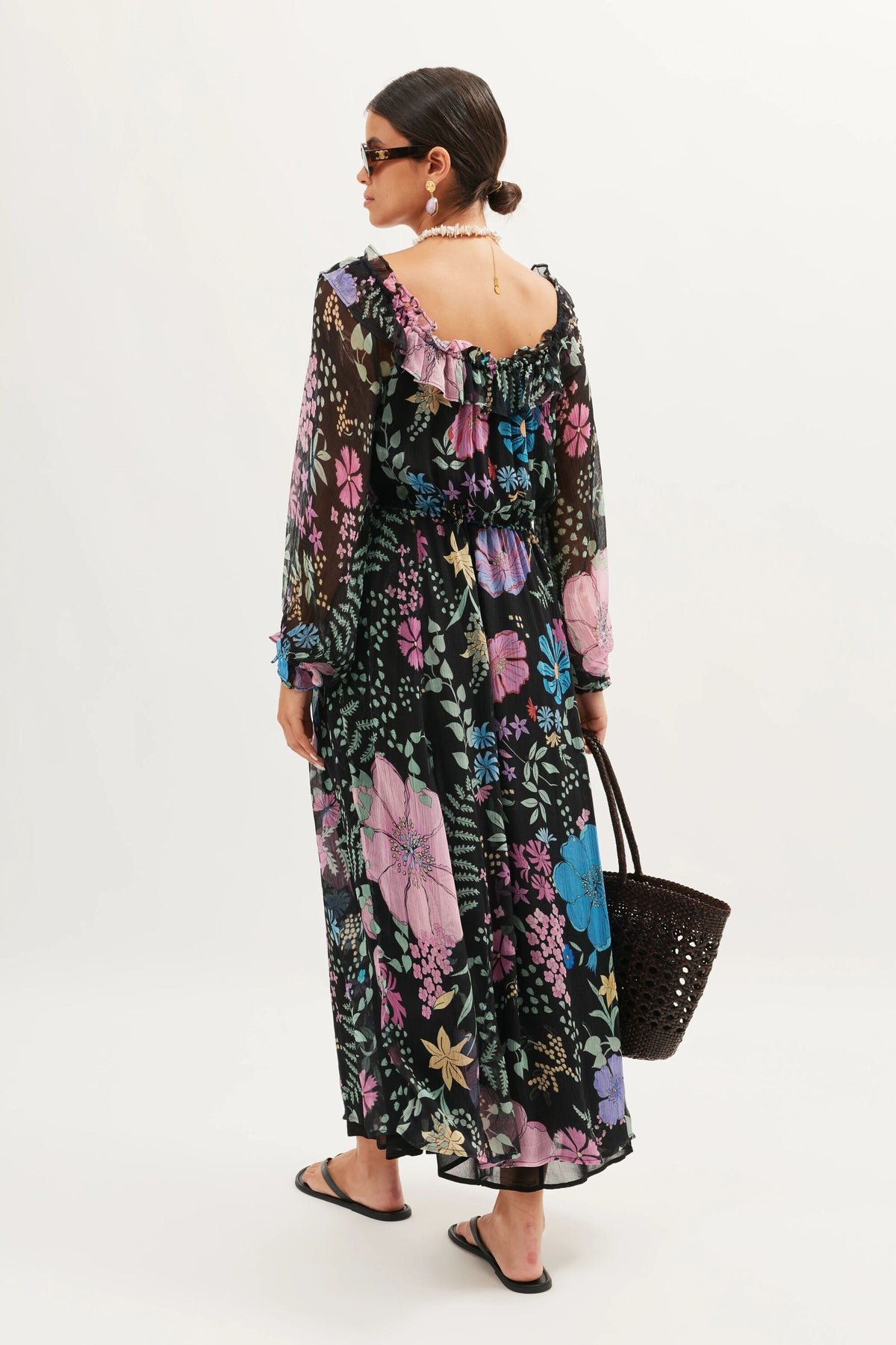 A woman faces away in the Alina – Bhatia Floral Black off-shoulder maxi dress with a vibrant floral print. She styles it with black sandals, sunglasses, a necklace, and a large black woven tote bag, her hair tied back.
