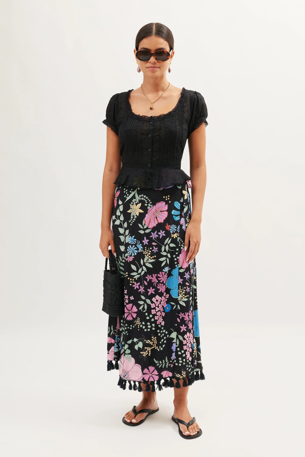 A woman wearing the Parisa - Bhatia Floral Black long skirt, a black short-sleeve blouse, black sandals, sunglasses, and holding a small black bag stands against a plain white background.