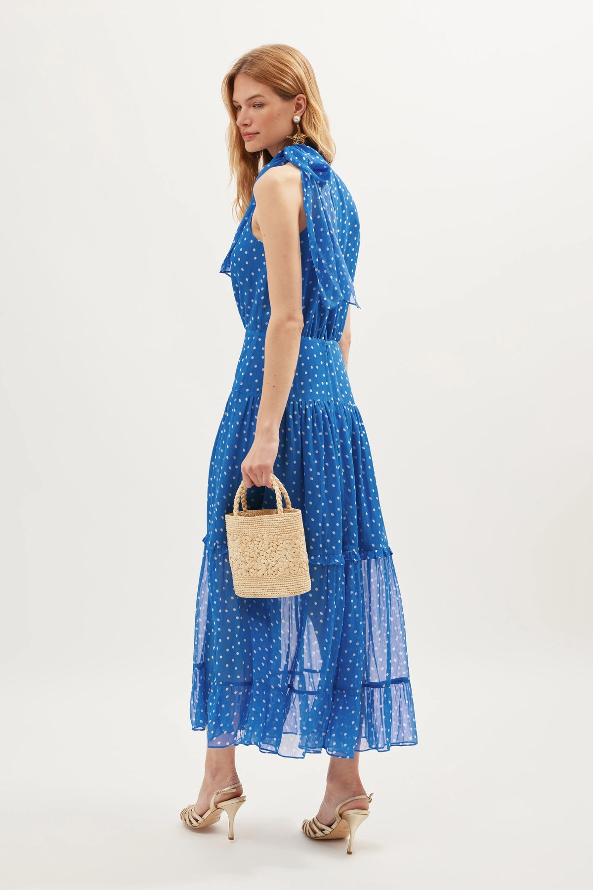 A woman in the Abi - Layla Dot Blue sleeveless polka dot dress with beige heeled sandals and a small straw handbag stands against a white background, looking over her shoulder—a chic occasionwear look.