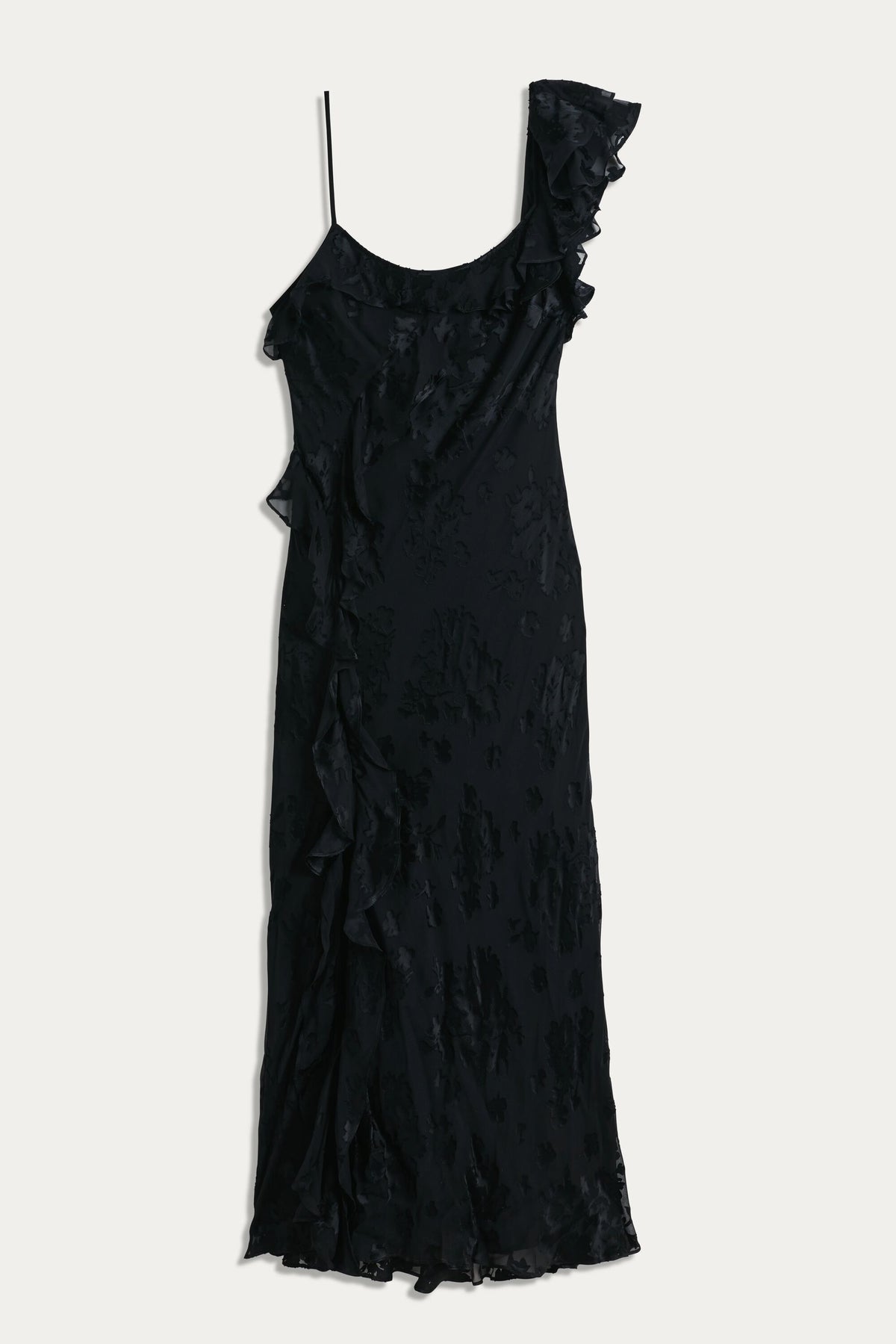 The Ester - Bouquet Devoré Black is a sleeveless black slip dress with one thin shoulder strap, a ruffled asymmetrical detail, cascading side ruffles, and a subtle floral pattern—ideal for an elegant evening look.