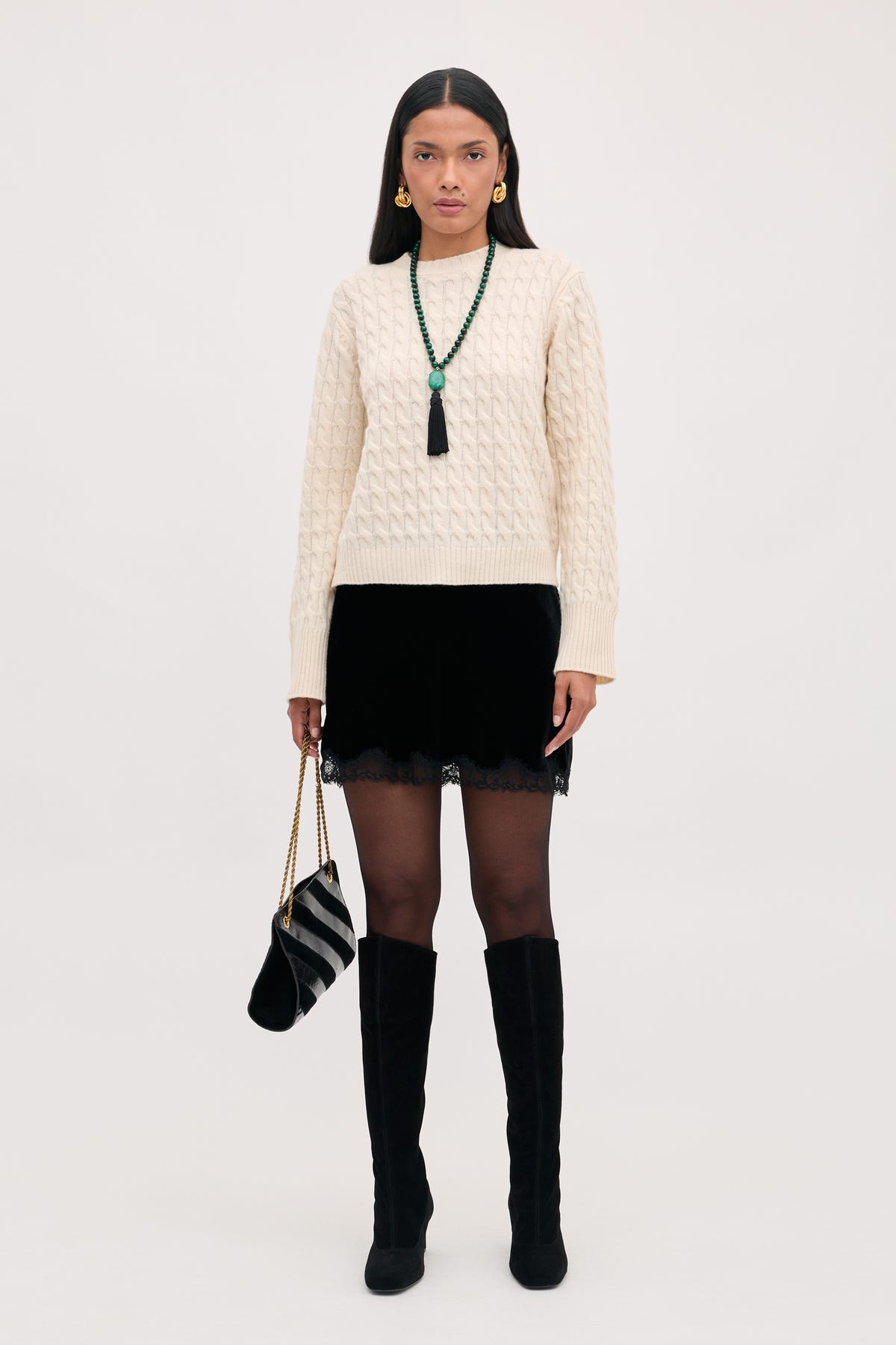 A model wears a cream cable-knit sweater, a black lace trim skirt, sheer tights, and black knee-high boots while carrying the Kornelia - Black handbag featuring a gold chain strap.