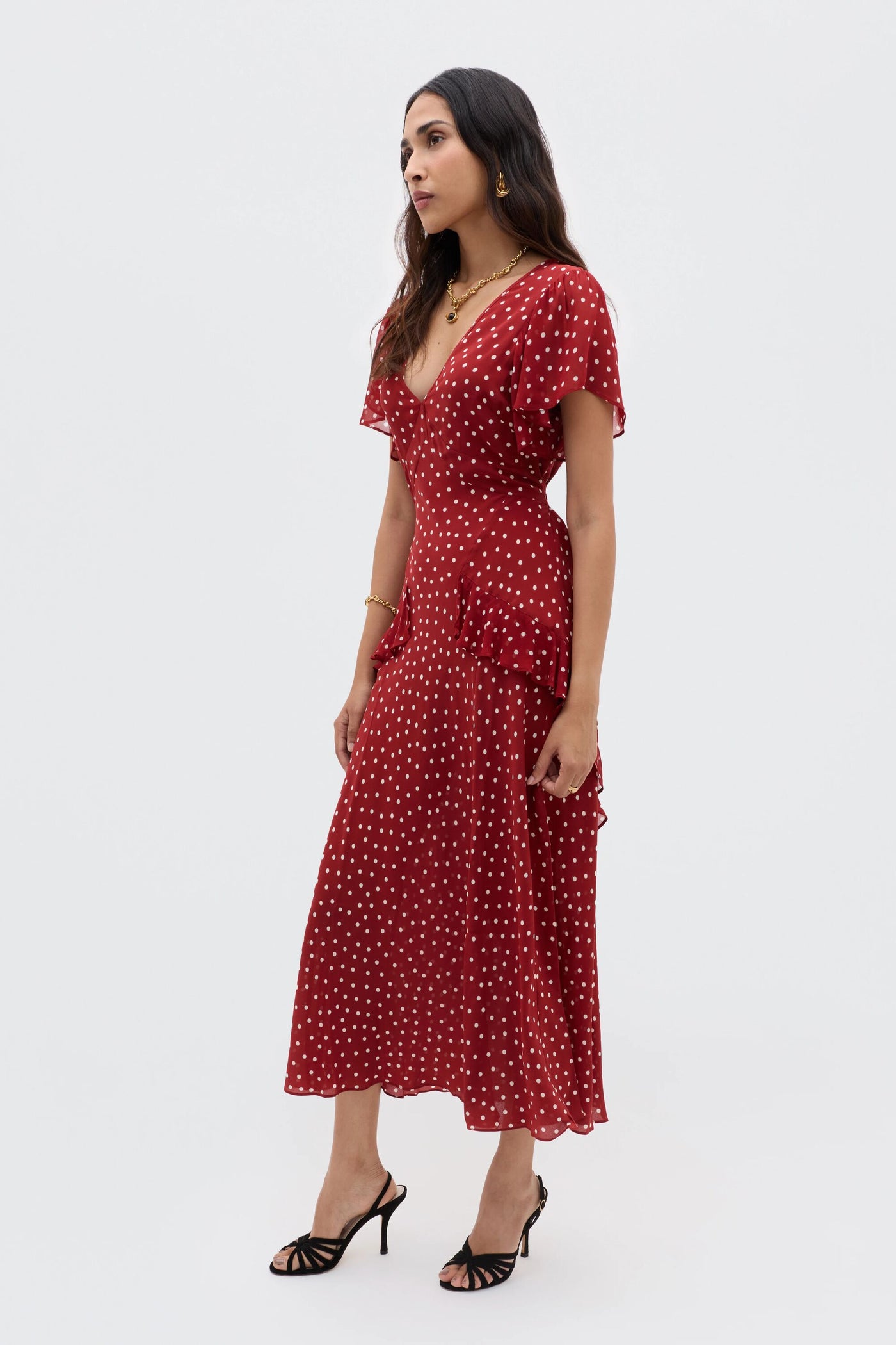 A woman in the RIXO Evie - Layla Dot Red dress with short sleeves, v-neck, and ruffle details stands in heeled sandals against a plain white background.