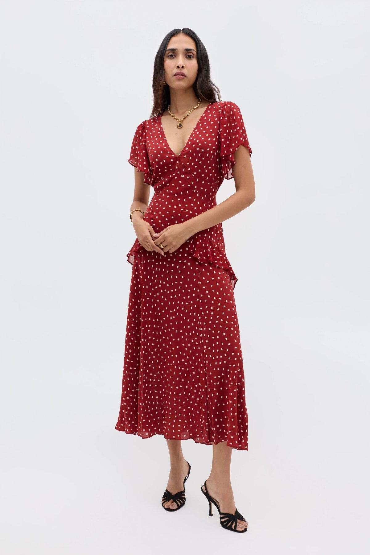 A woman wears the Evie - Layla Dot Red dress—red, short-sleeved, V-neck with polka dots—paired with black heeled sandals, standing against a plain light gray background.