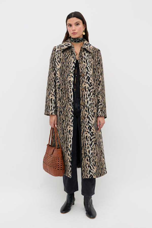 A woman stands against a white background wearing the Milly - Bohemia Leopard mid-length coat over dark clothing, paired with black ankle boots, a patterned scarf, gold earrings, and carrying a brown woven handbag as she looks at the camera.