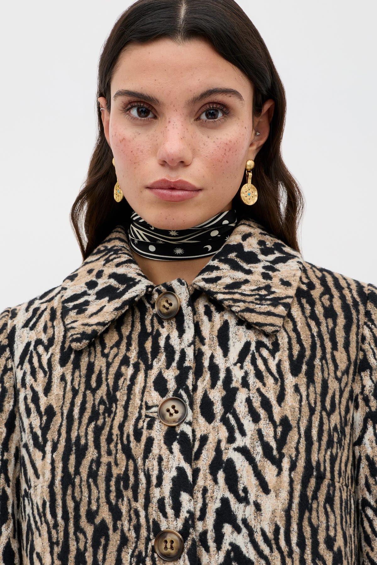 A woman with long dark hair and freckles wears the Milly - Bohemia Leopard mid-length coat with large buttons, gold drop earrings, and a black-and-white scarf around her neck, standing against a plain light background.
