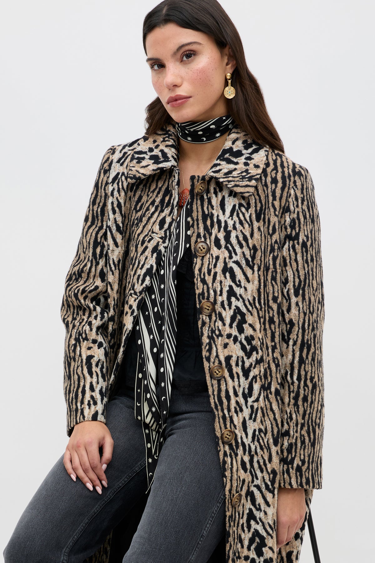 A woman wearing the Milly - Bohemia Leopard mid-length coat, black jeans, a patterned black and white scarf, and gold earrings sits facing the camera against a plain white background.