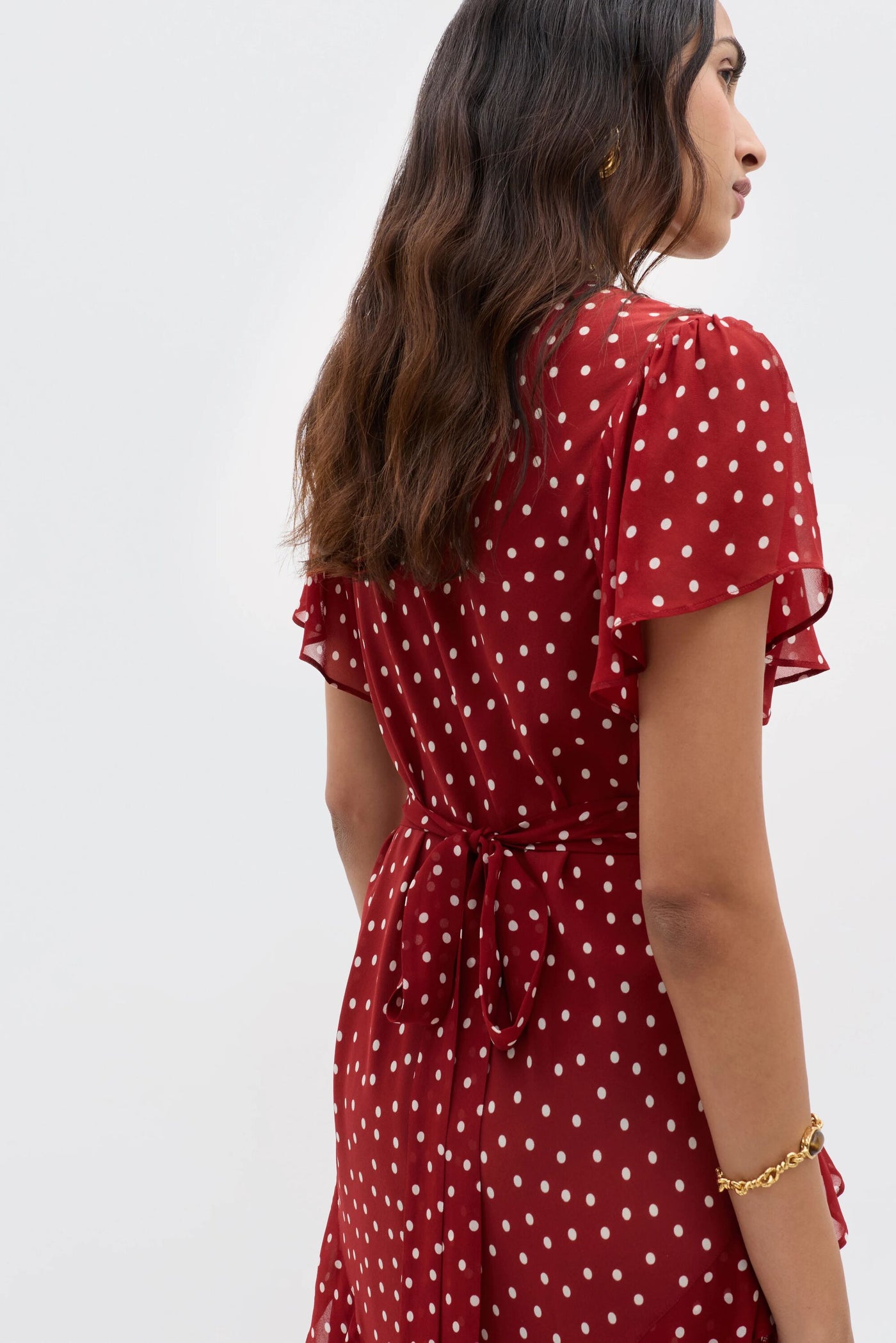 A woman with wavy brown hair wears the RIXO Evie - Layla Dot Red dress, featuring short flutter sleeves and a waist tie, standing with her back slightly turned against a plain light background.