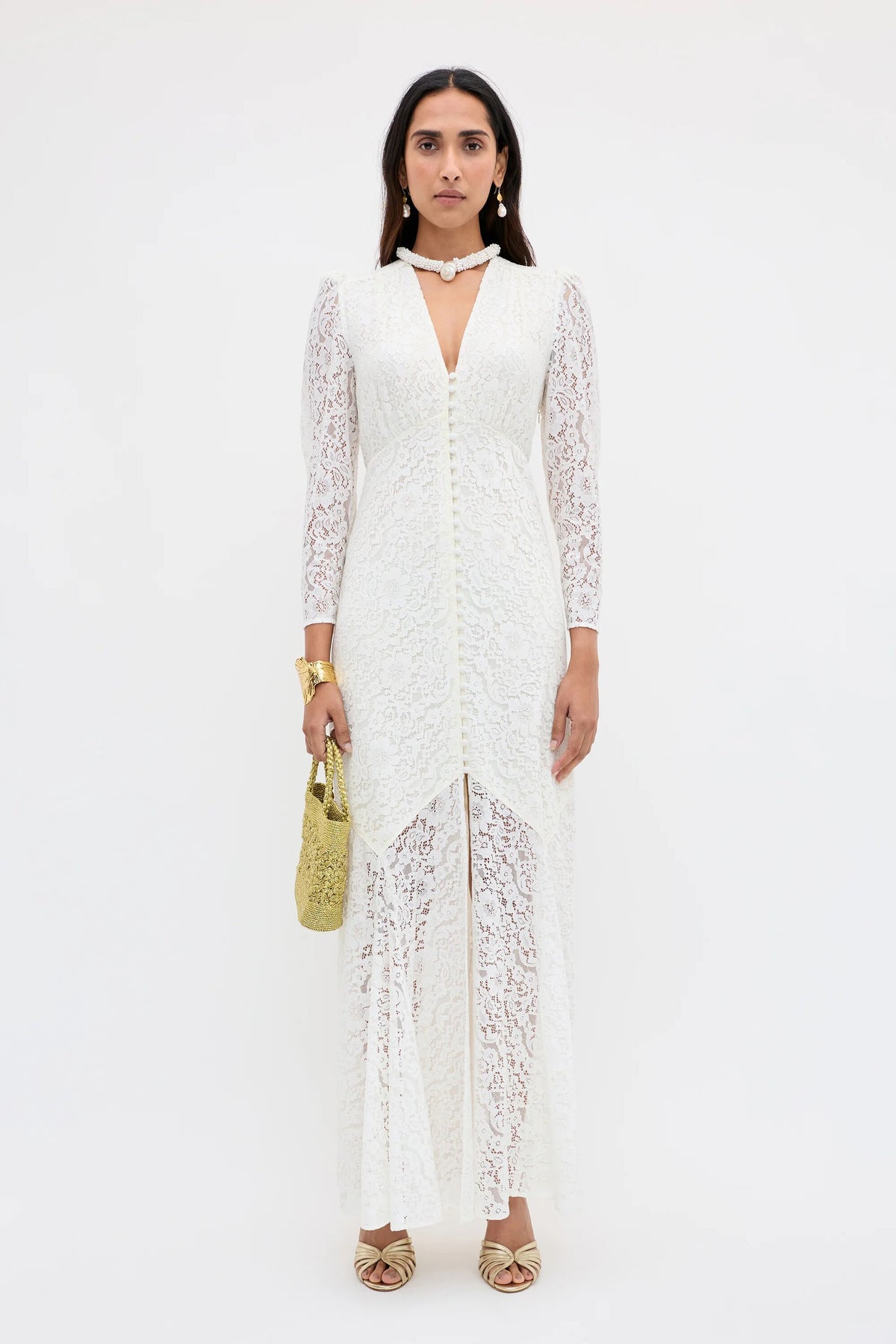 A woman poses against a white background wearing the Fleur - Ivory, a long-sleeve, floor-length corded lace dress with a deep V neckline. She accessorizes with a small gold handbag, gold sandals, statement jewelry, and straight dark hair.