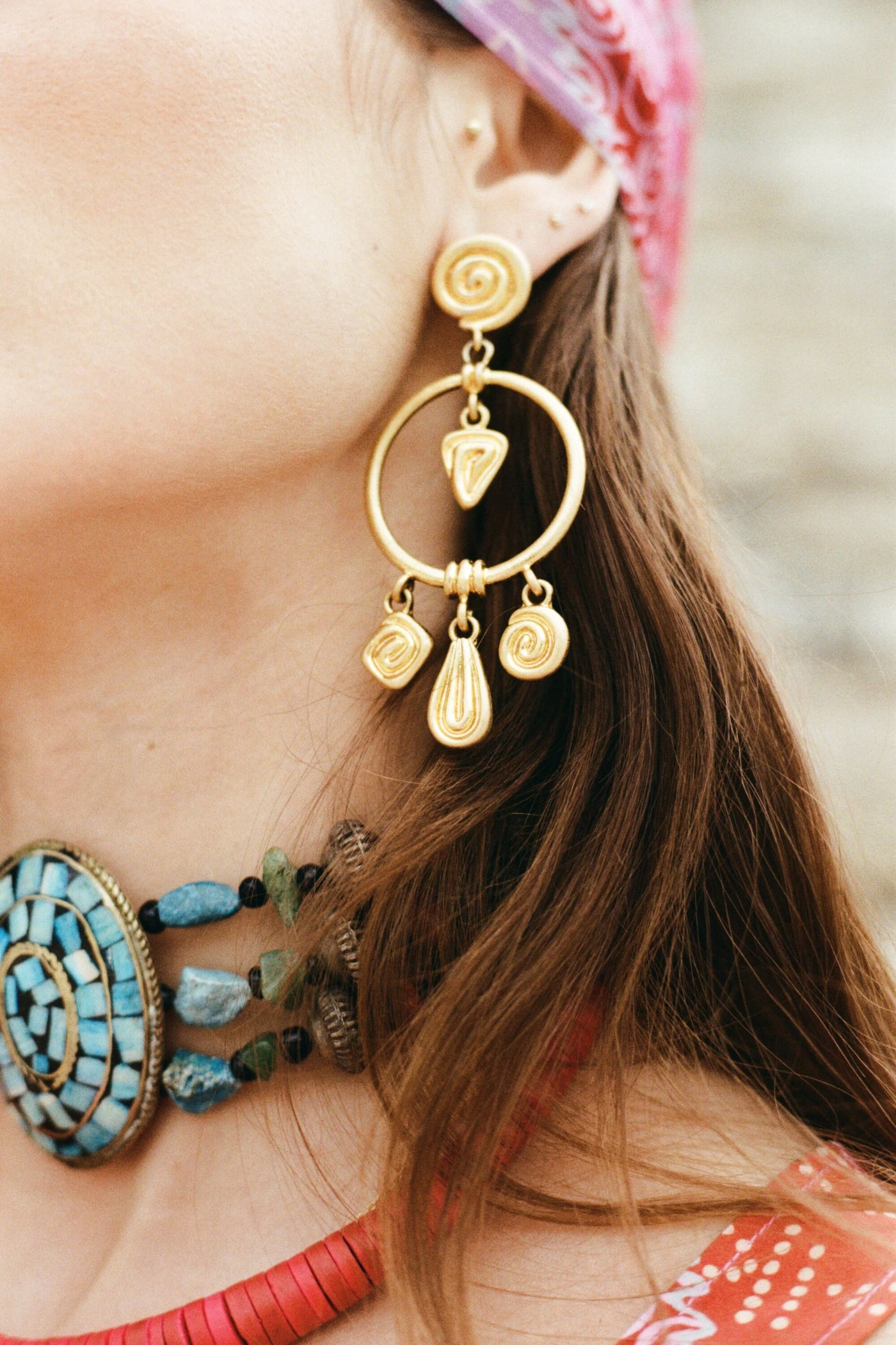 Close-up of a woman wearing Thome - Gold drop earrings and an ornate turquoise bead necklace. Her brown hair is down, with a pink patterned headscarf. Only part of her face is visible, showcasing her unique bohemian jewelry style.
