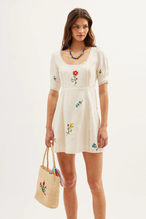 A woman poses against a white background in the Marne – Ombre Floral Embroidery Cream dress, accessorized with a straw tote bag featuring colorful accents and a blue beaded necklace, her long hair loose for an effortlessly chic look.