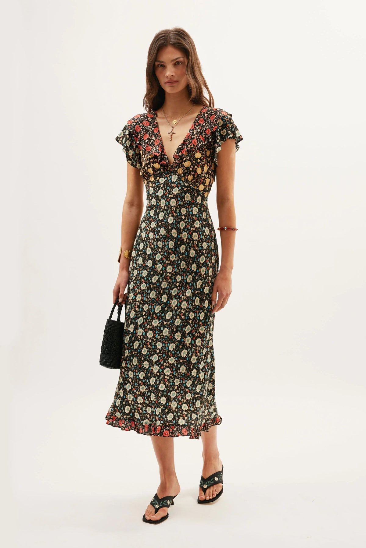 A woman stands against a plain background wearing the Bethea - Eva Floral Mix Black dress. She pairs it with a small black handbag, black sandals, gold jewelry, and has loose brown hair.