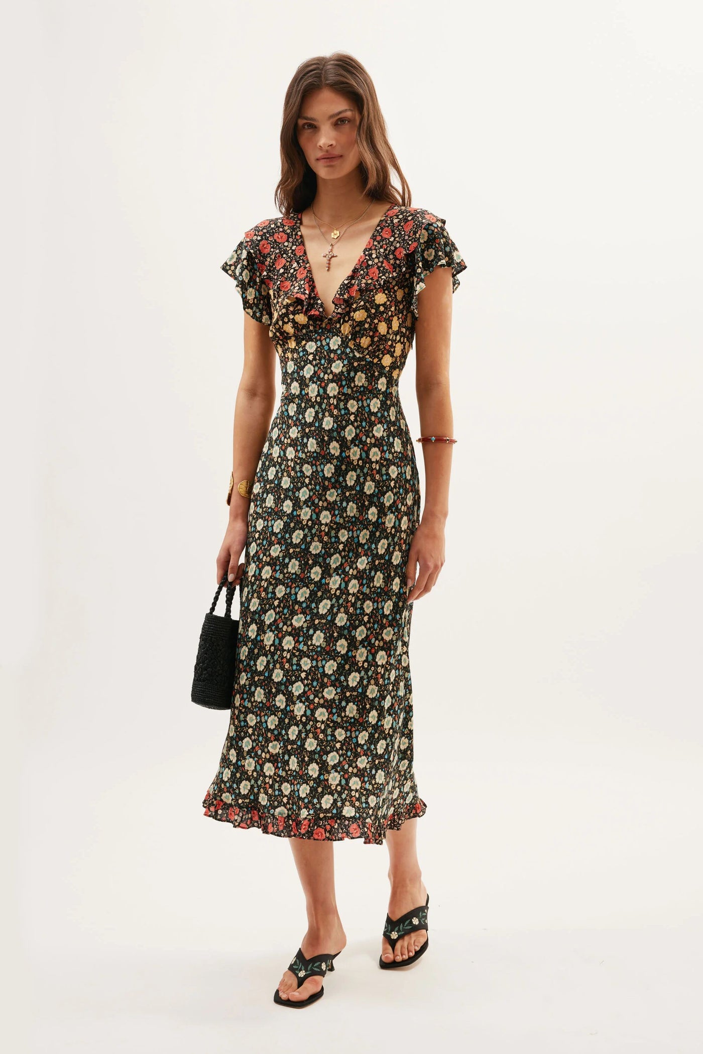 A woman stands against a plain background wearing the Bethea - Eva Floral Mix Black dress. She pairs it with a small black handbag, black sandals, gold jewelry, and has loose brown hair.