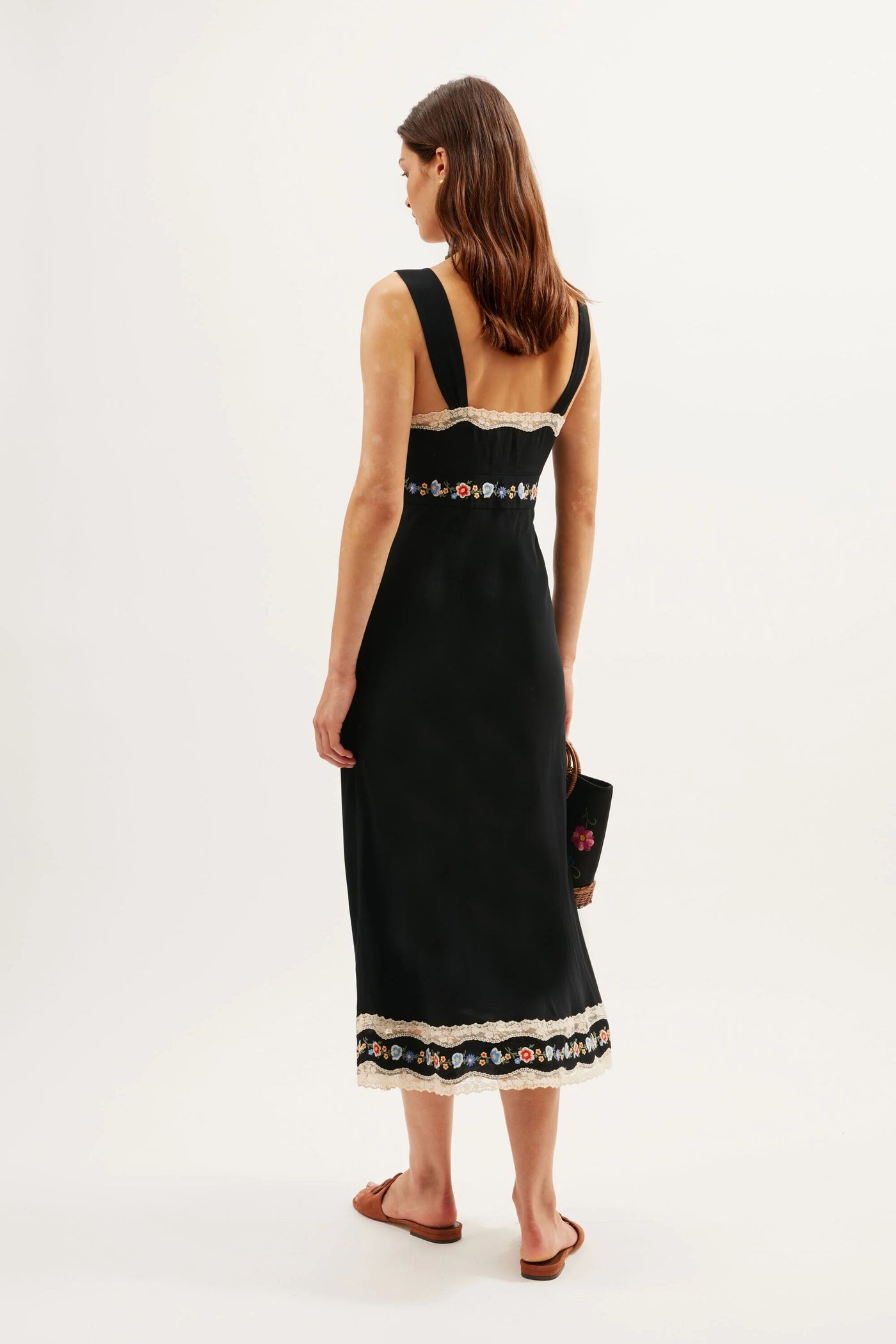 A woman with straight brown hair faces away, wearing the Lucie - Bloom Embroidery Black sleeveless crepe de chine dress with lace trim and holding a black clutch. She pairs brown flat sandals against a plain white background.