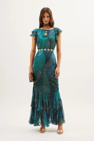 A woman stands against a plain background wearing the Ordinia – Ibiza Night Teal, a flowing formal dress with ruffled sleeves and hem, accessorized with gold jewelry, open-toe heels, and a clutch—ideal for special events.