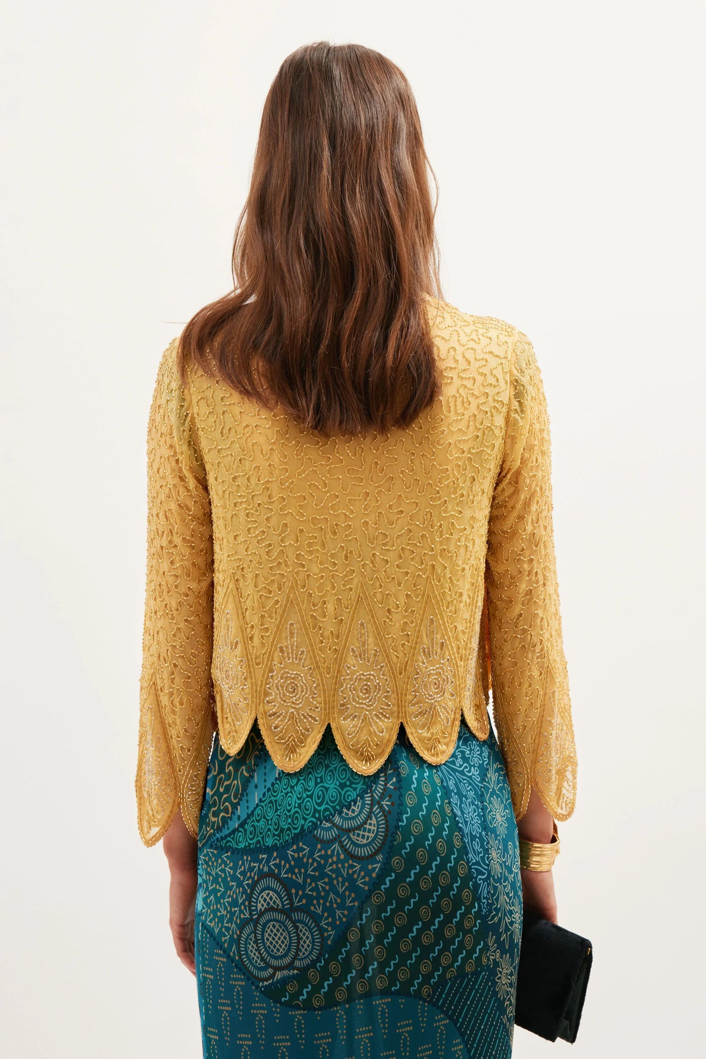 A woman with wavy brown hair faces away, wearing the Mateo Florence Embellishment Gold top—featuring gold embroidery and scalloped edges—paired with a teal patterned skirt and holding a black clutch against a plain light background.