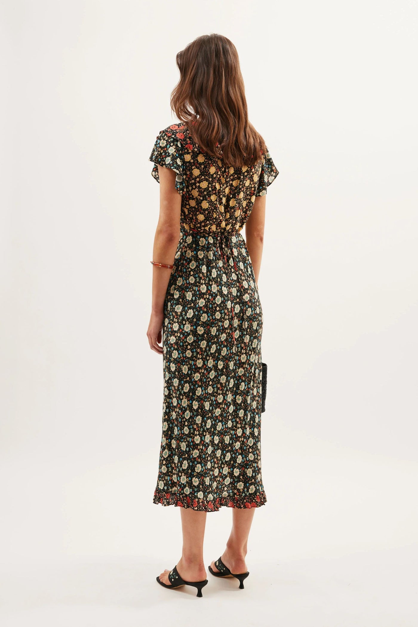 A woman with wavy brown hair wears the Bethea - Eva Floral Mix Black dress, featuring a black crepe fabric with a colorful floral print, short flutter sleeves, and a ruffled neckline. She holds a black clutch and wears mid-heel sandals.