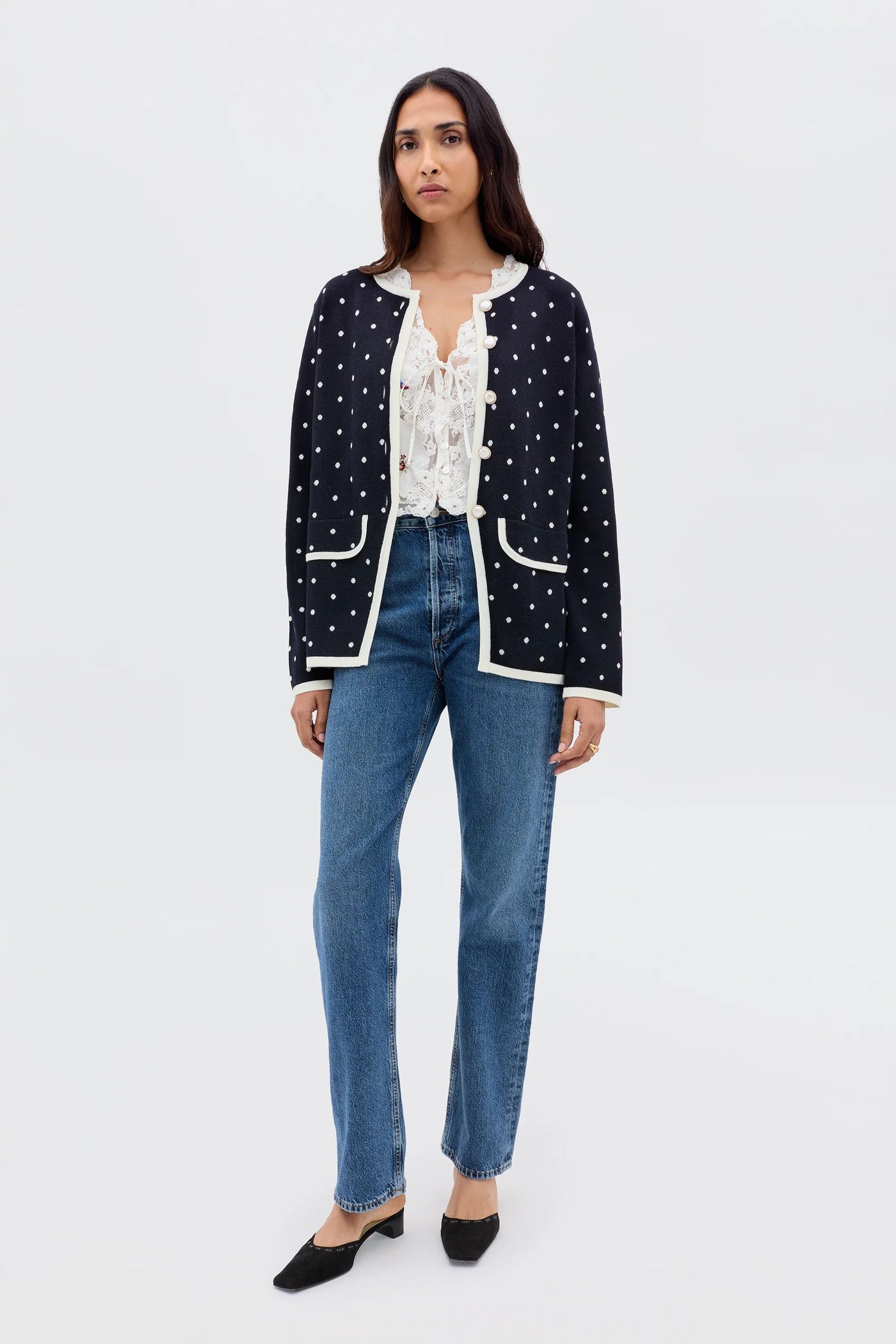 Woman standing against a plain background, wearing the Mirja - City Spot Jacquard Navy cardigan over a frilled white blouse, blue jeans, and black heeled shoes.