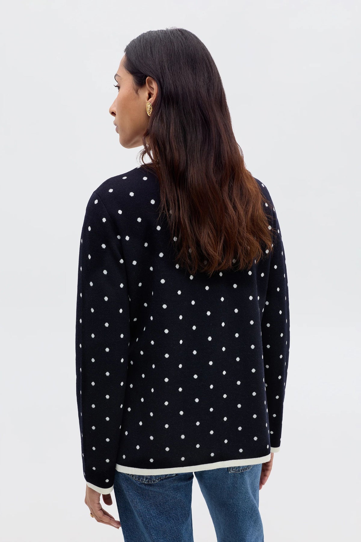 A woman with long brown hair faces away, wearing the Mirja - City Spot Jacquard Navy cardigan with white polka dots and blue jeans, standing against a plain light background.