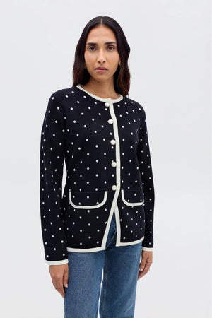 A woman wears the Mirja - City Spot Jacquard Navy, a navy merino wool cardigan with white spots, cream trim, and large buttons, paired with blue jeans against a plain light background.