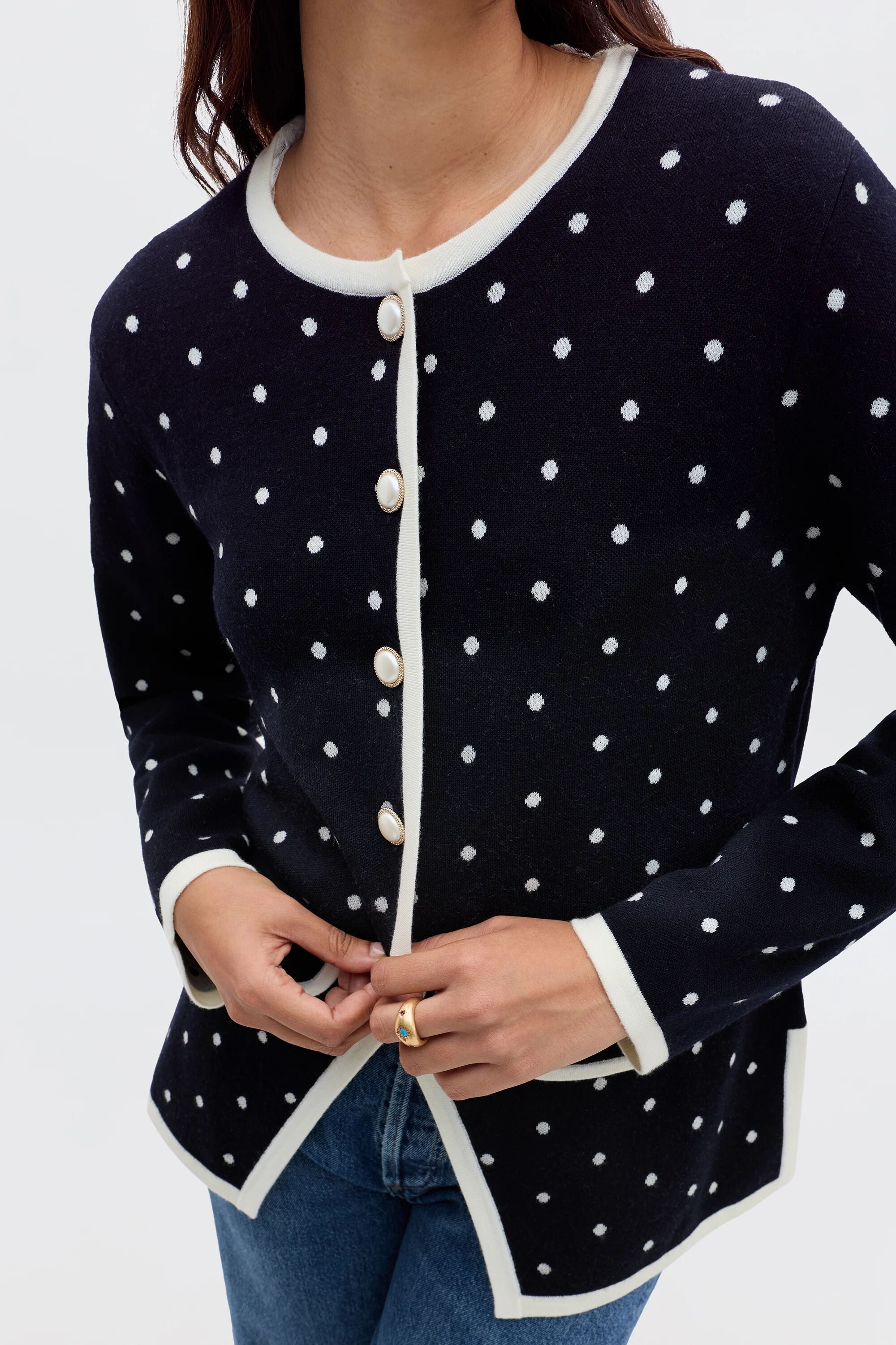 Someone wearing the Mirja - City Spot Jacquard Navy cardigan, a navy blue piece with white polka dots and trim, paired with blue jeans, adjusts a button near the hem.