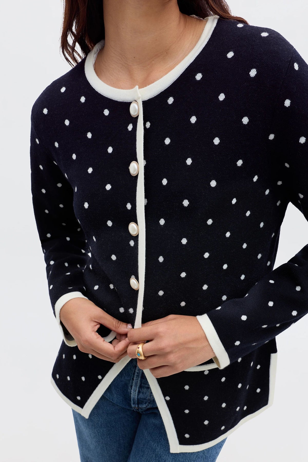 Someone wearing the Mirja - City Spot Jacquard Navy cardigan, a navy blue piece with white polka dots and trim, paired with blue jeans, adjusts a button near the hem.