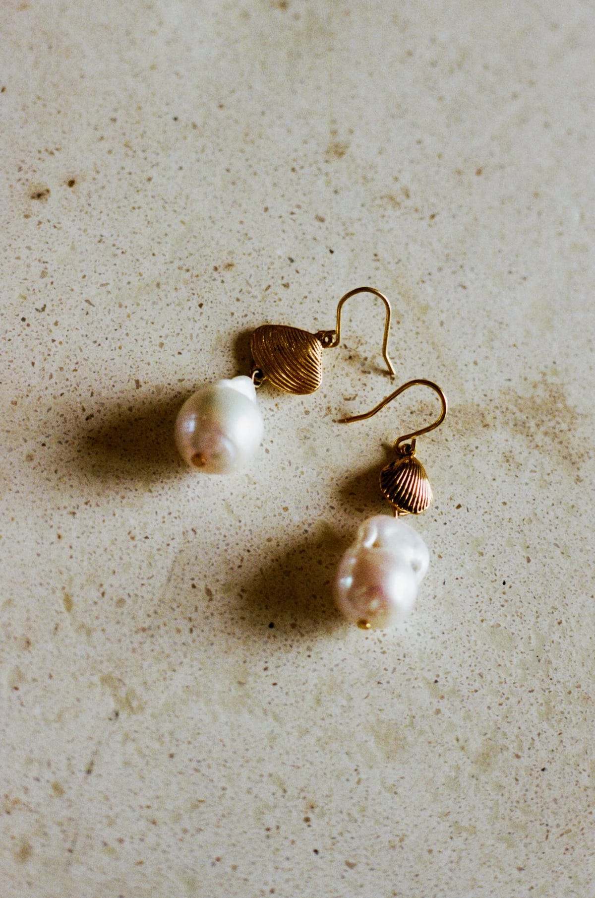Rosaria - Gold features gold-plated drop earrings with textured shell-shaped accents and large, irregular white pearls, displayed on a speckled beige surface.