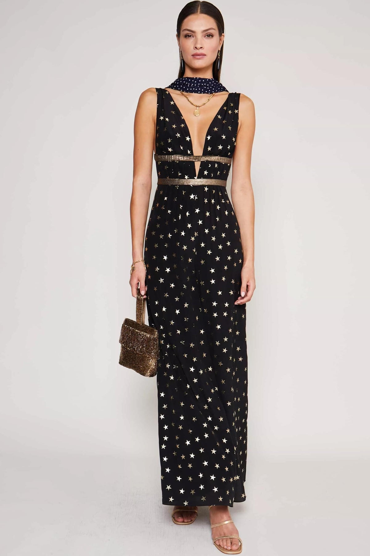 A woman stands facing forward in the Hollywood - Star Foil sleeveless black gown with a deep V-neck and gold-foil star print, accessorized with a glittery handbag, strappy sandals, layered necklaces, and a dark dotted scarf—party ready.