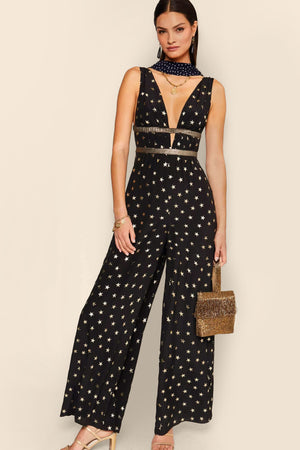 A party-ready woman in the Hollywood - Star Foil jumpsuit, featuring a black base with gold-foil star print.