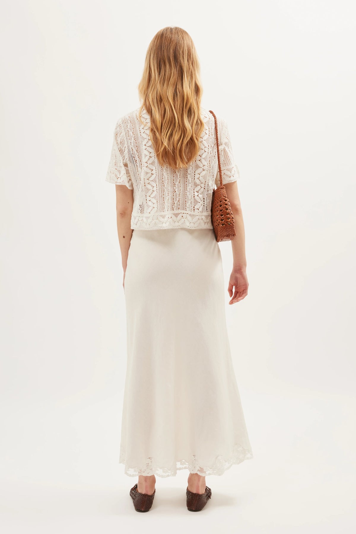 A woman with long blonde hair stands facing away, wearing the Diona - Ivory vintage lace short-sleeve top with a boxy shape and a long white skirt. She carries a brown woven shoulder bag and wears brown shoes against a plain white background.