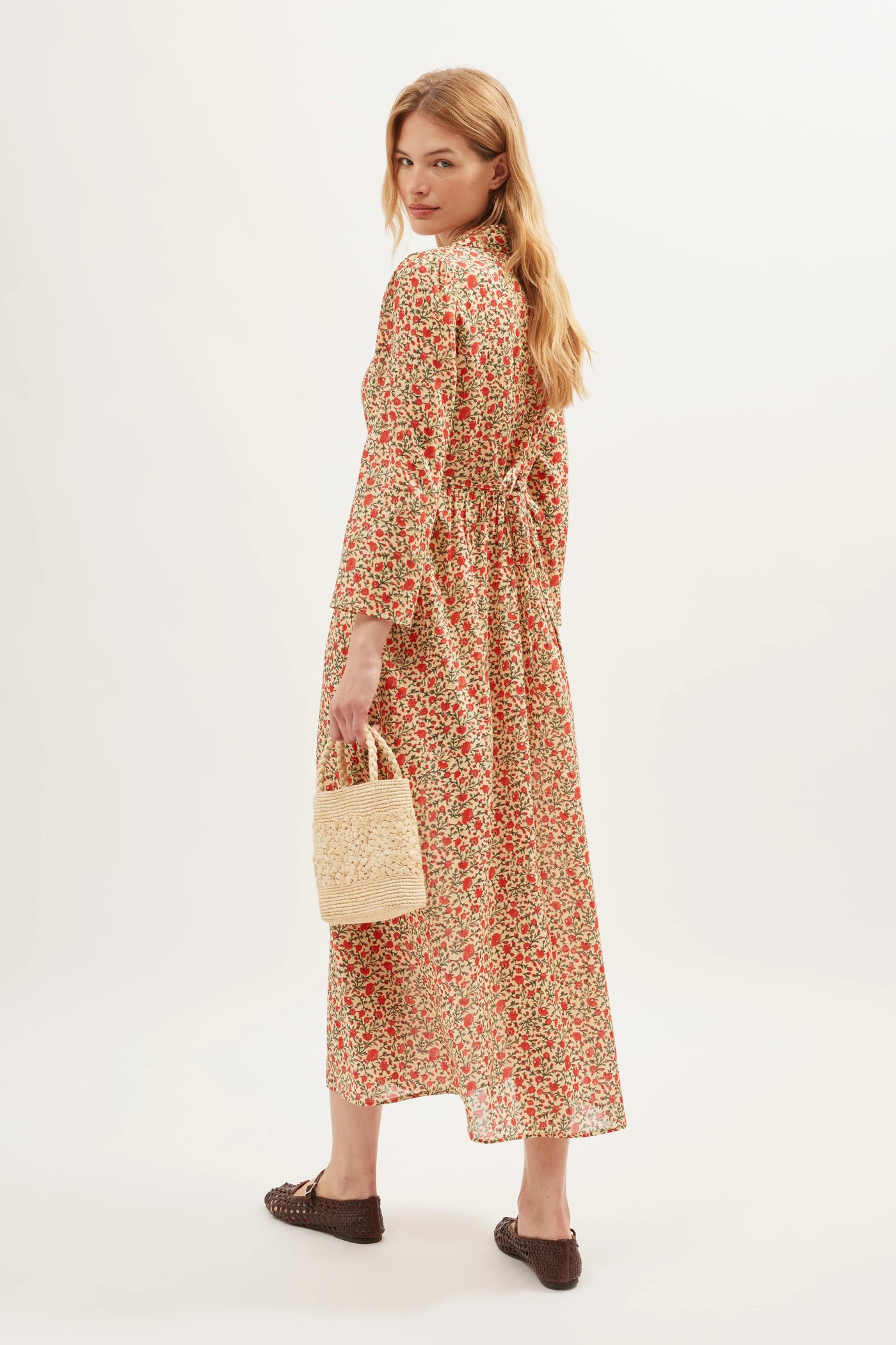 A woman with long blonde hair wears the Franchi - Ditsy Trail Cream dress, featuring red and white patterns. She carries a woven beige handbag and stands in brown flats, effortlessly posing against a light background.