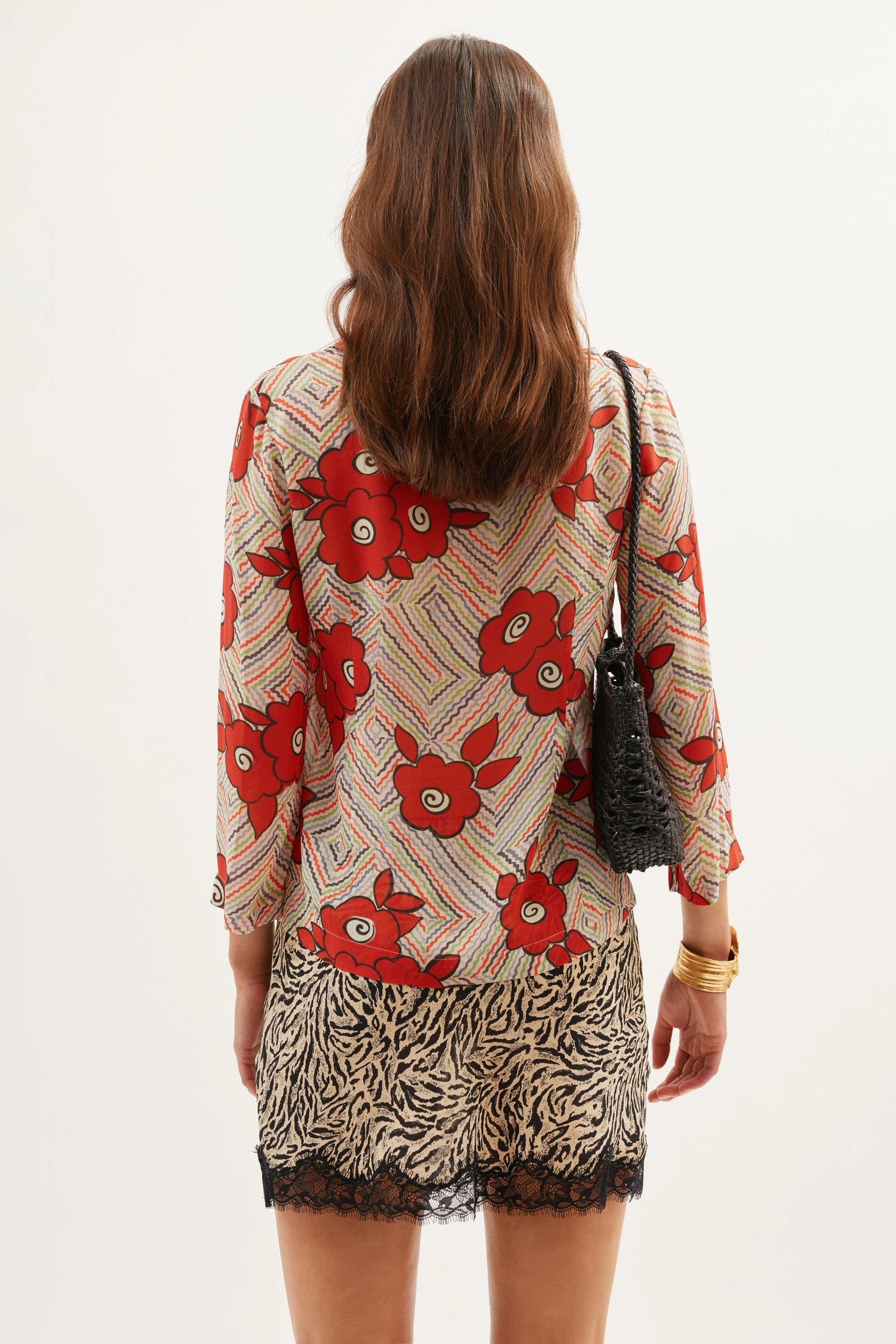 A woman with wavy brown hair faces away, wearing the Henny - Milos Floral Cream top, featuring red flowers and patterned stripes, a zebra-print skirt with lace trim, a black shoulder bag, and gold bangles.