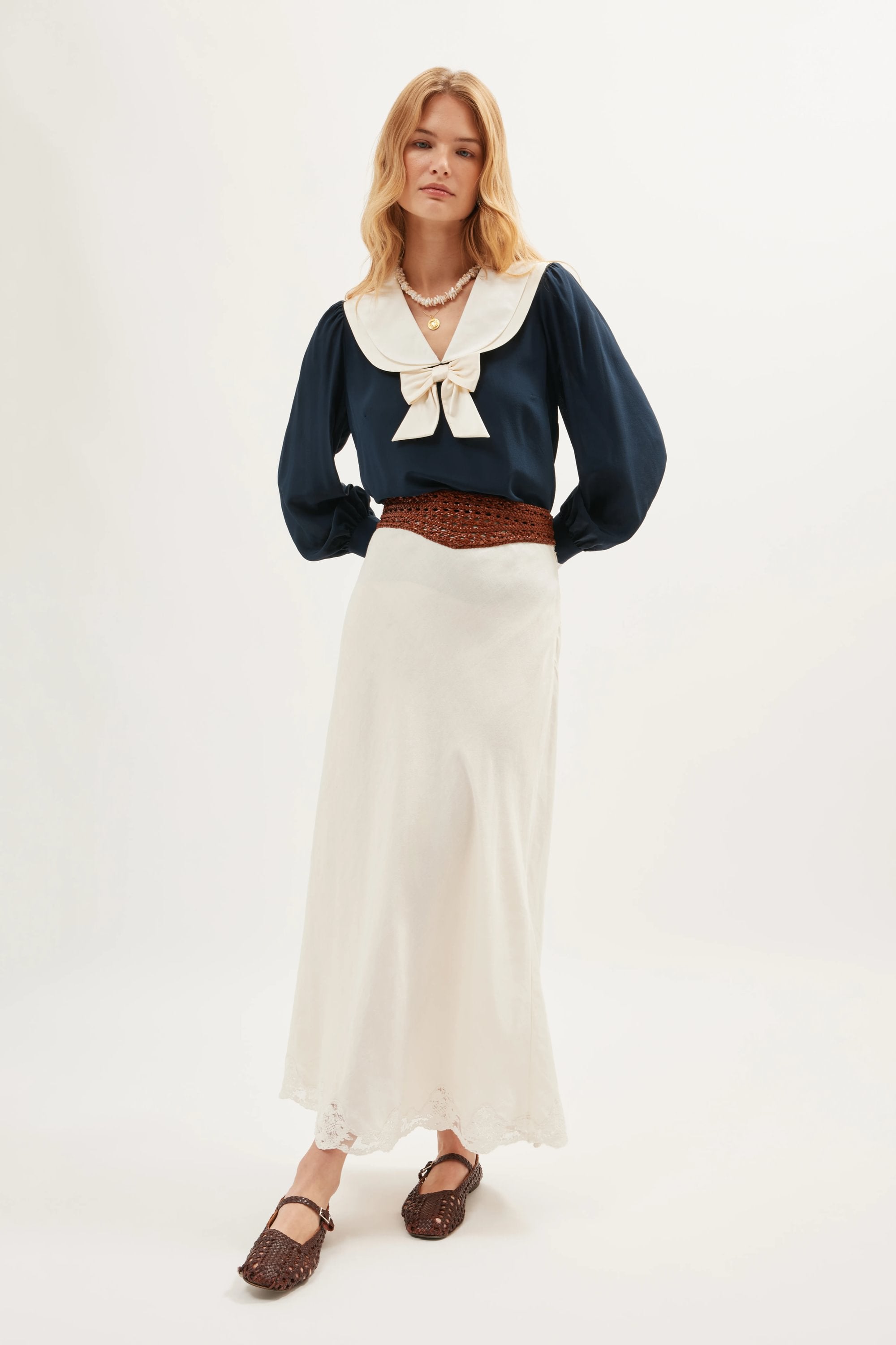 A person wears the Crystal - Cream, a long ivory linen skirt, paired with a navy long-sleeve blouse with a large white collar and bow, brown woven belt and shoes, and layered necklaces, standing against a plain white background.