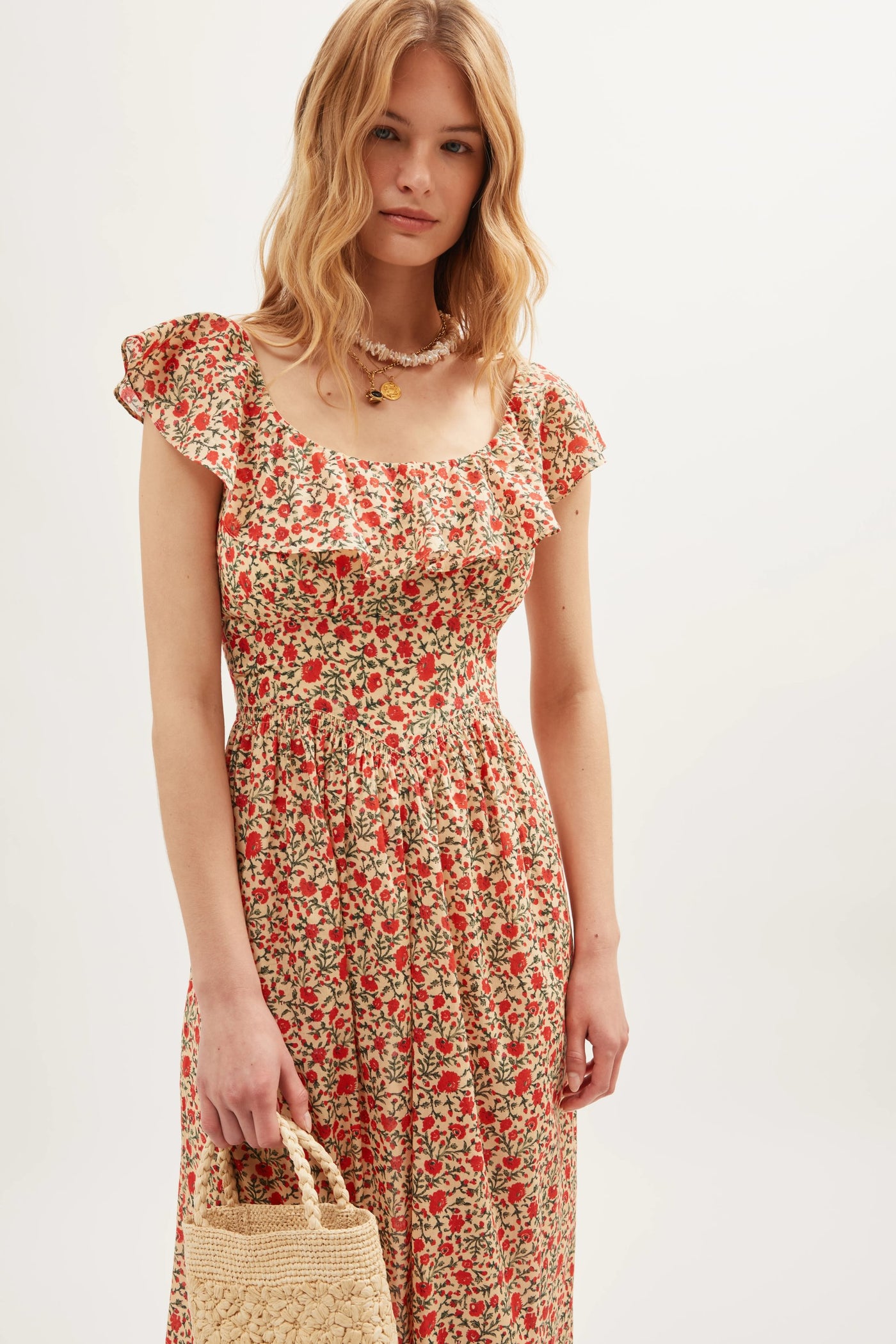 A woman with wavy blonde hair wears the "Ebony - Ditsy Trail Cream" floral dress with ruffled sleeves and holds a woven straw bag. She accessorizes with a necklace, standing against a plain, light-colored background.