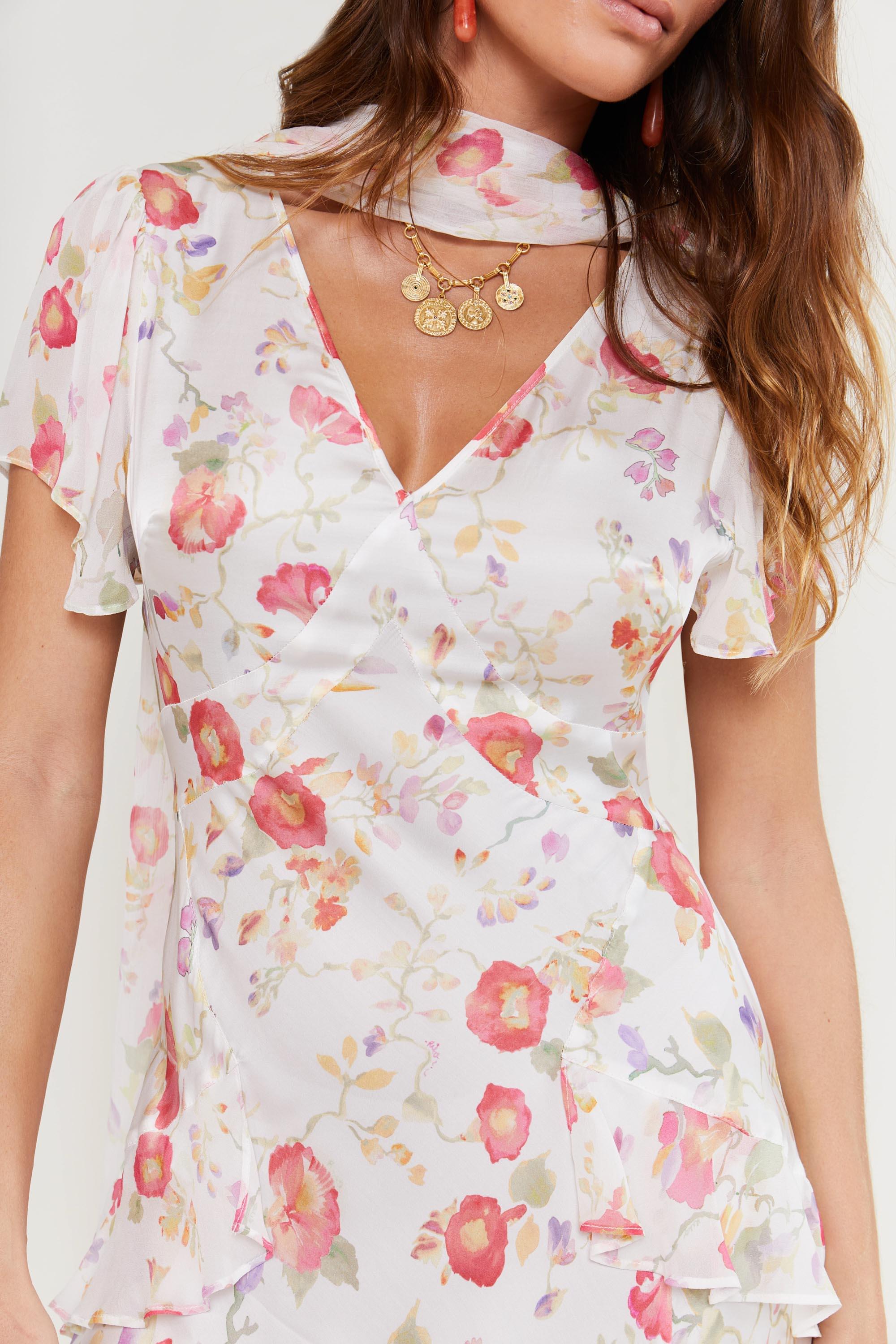 A woman wears the "evie - waterblossom ivory" dress—a white Hand painted floral piece with pink and orange flowers, layered short sleeves, and matching scarf. She has long wavy brown hair, gold necklaces, and orange earrings; only her upper body is visible.