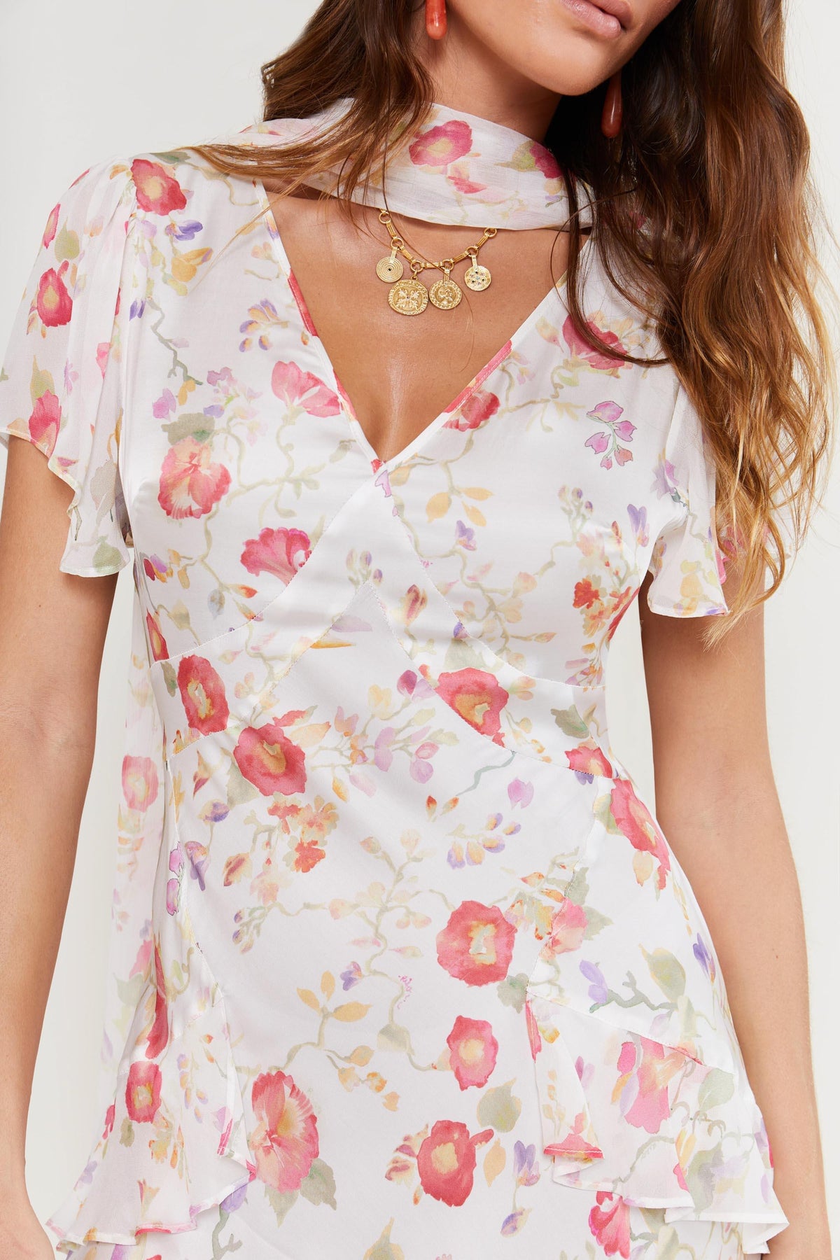A woman wears the "evie - waterblossom ivory" dress—a white Hand painted floral piece with pink and orange flowers, layered short sleeves, and matching scarf. She has long wavy brown hair, gold necklaces, and orange earrings; only her upper body is visible.