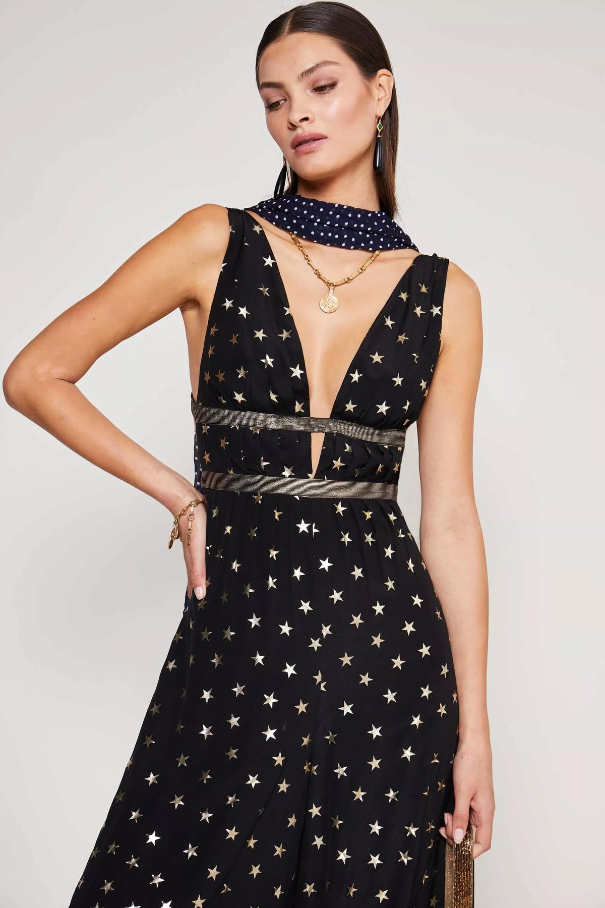 A woman wears the Hollywood - Star Foil sleeveless black dress with a gold-foil star print, deep V-neck, and black waistband. She accessorizes with layered necklaces, earrings, bracelets, and a navy polka-dot scarf.