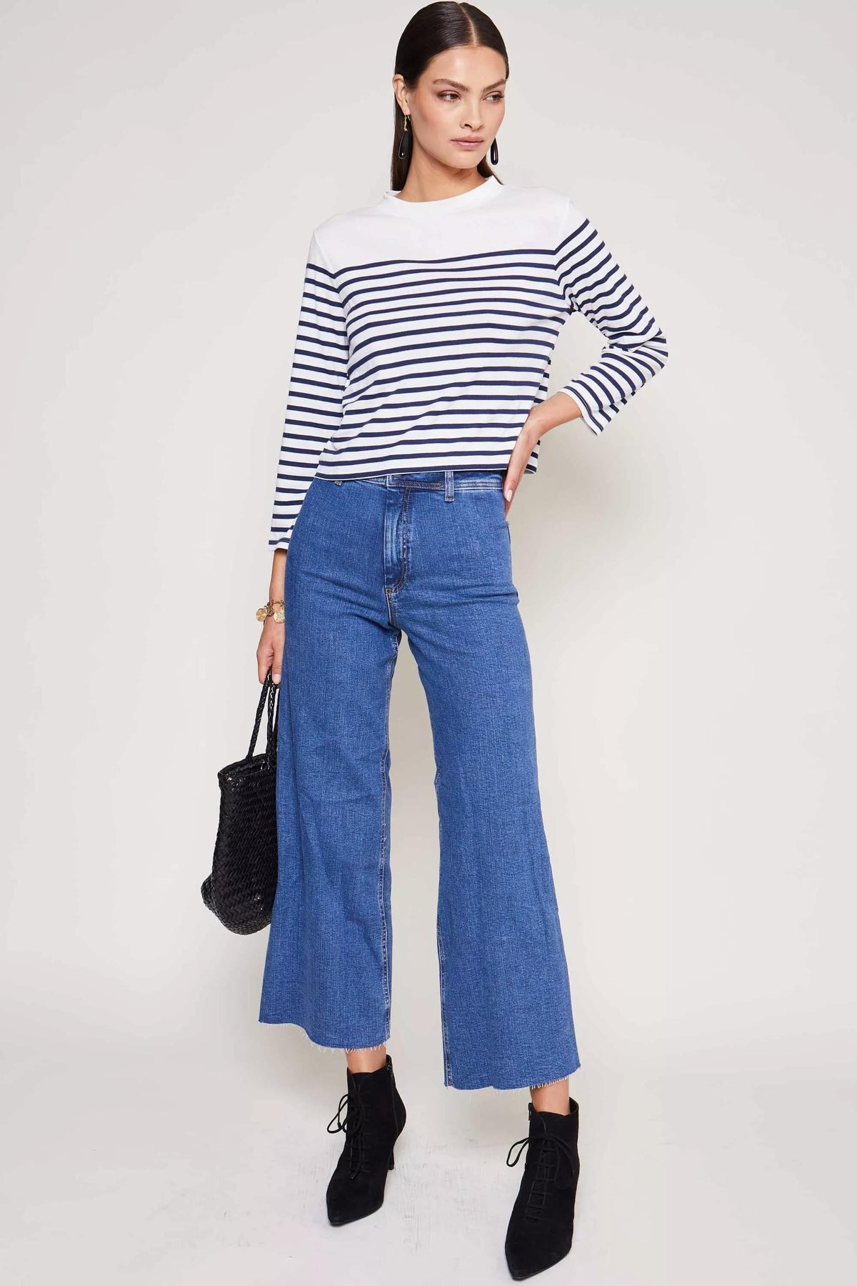 A woman stands against a plain background wearing the Francesco - Nautical Stripe Cream top, high-waisted wide-leg blue jeans, black ankle boots, and holding a black bag—a true wardrobe staple.